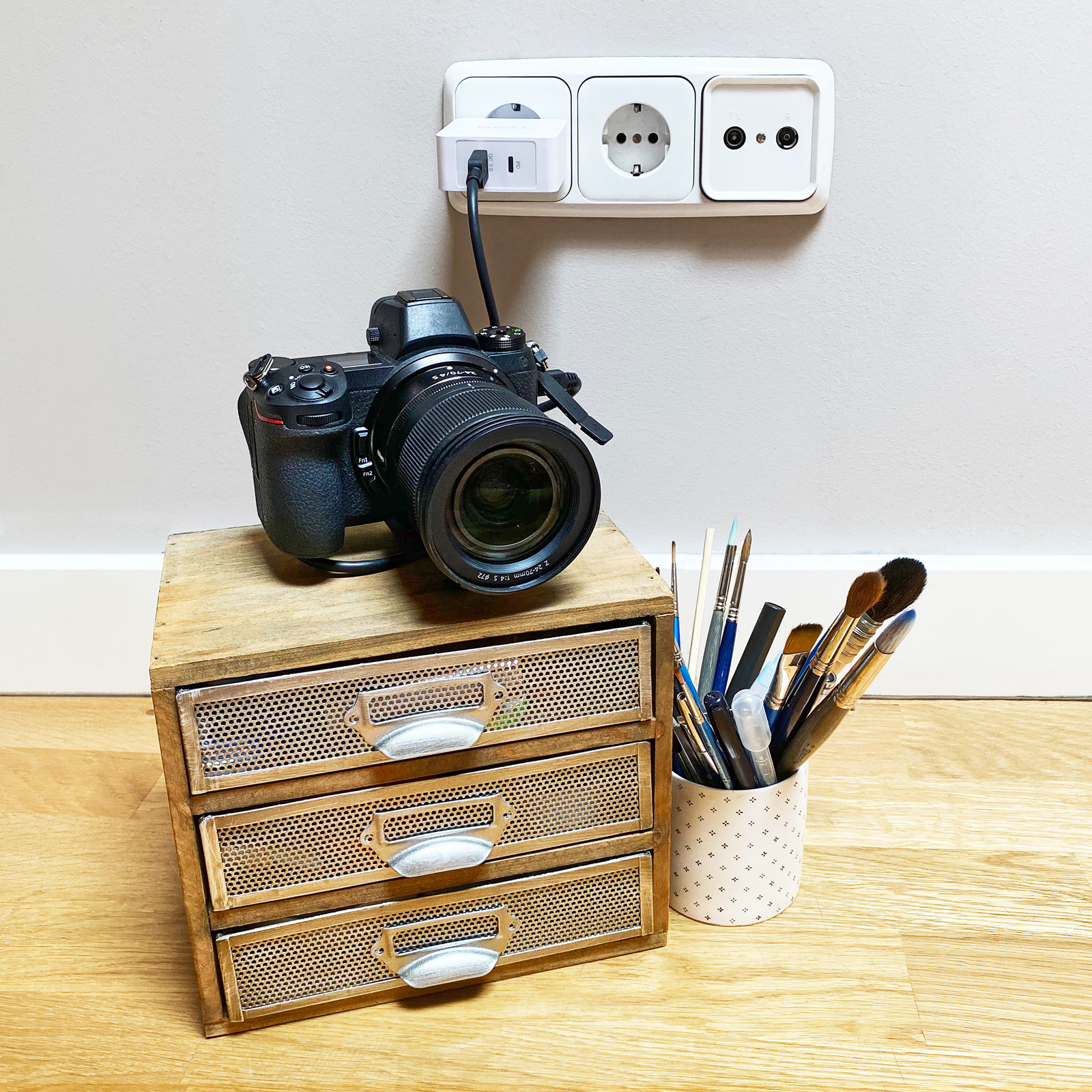 Una cámara en un cajón de madera, con pinceles en una taza y un enchufe en la pared.