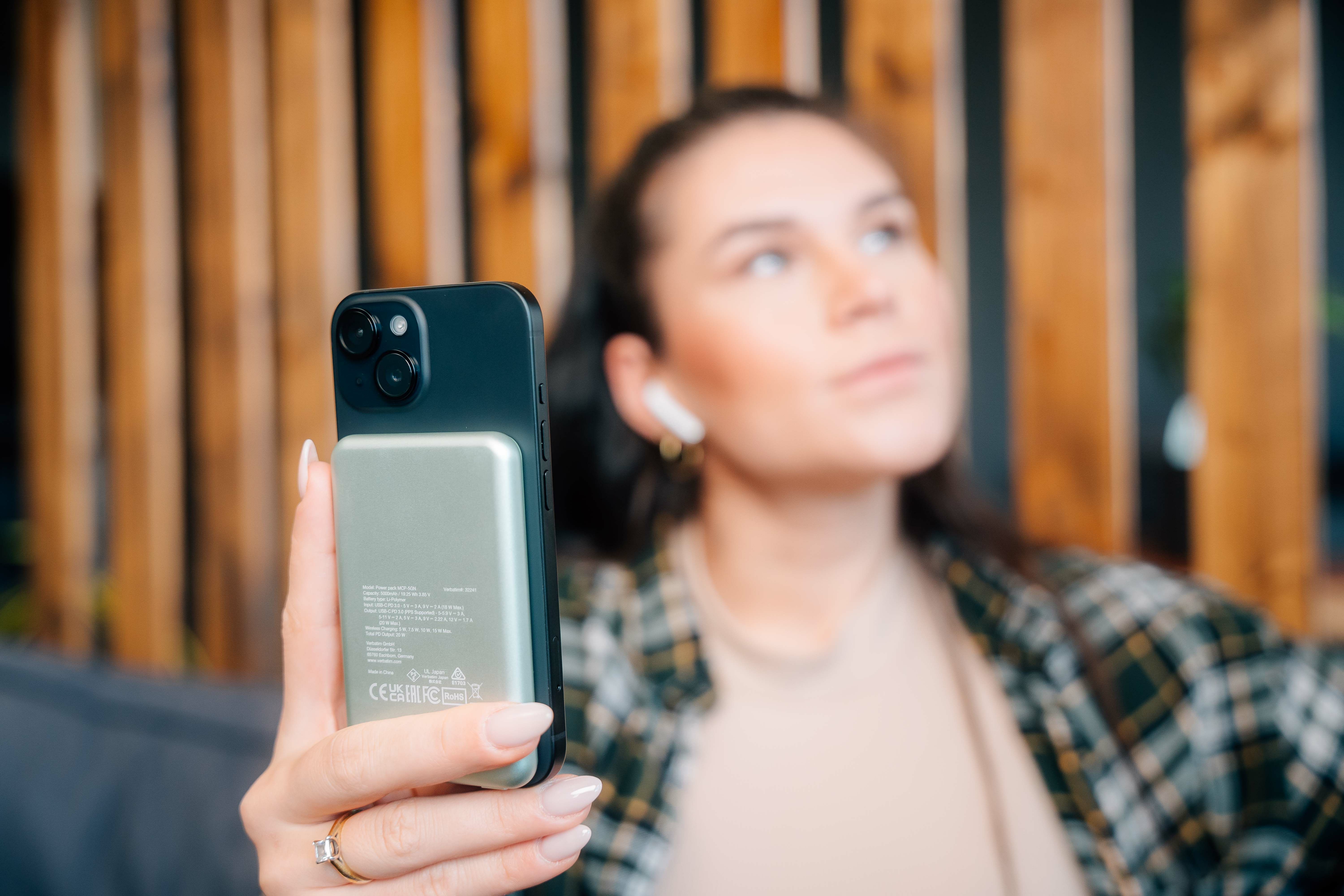 Een vrouw houdt een zwarte telefoon met een zilveren achterkant vast. Ze draagt oortelefoons en een ring. Achtergrond is wazig.