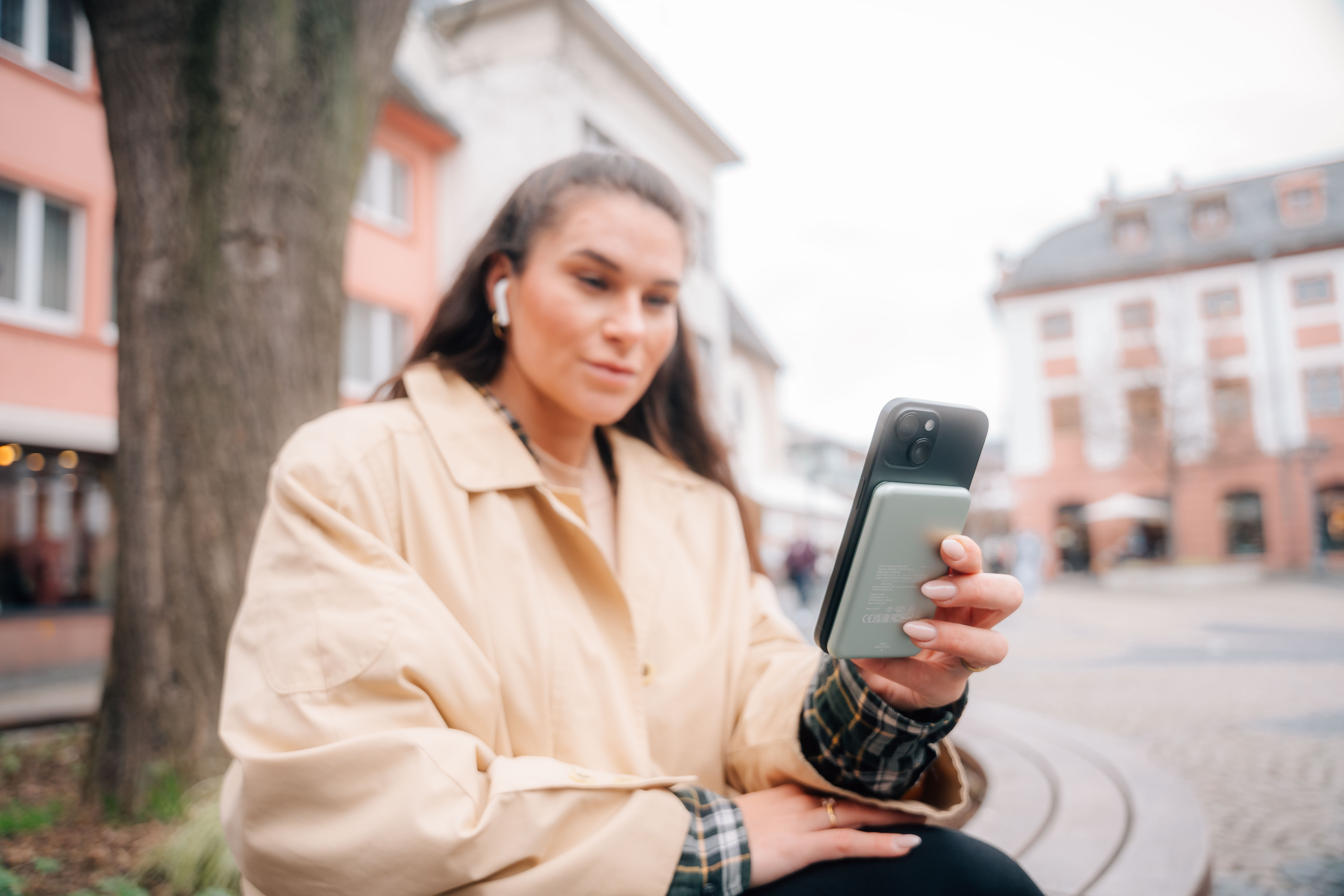 Vrouw houdt telefoon met powerbank vast, draagt beige jas, in stedelijke omgeving.