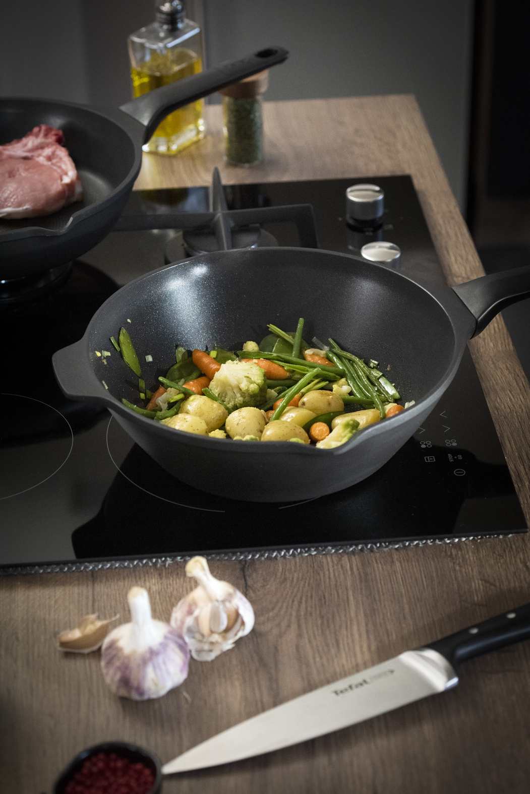 Een wok met groenten staat op een zwart fornuis. Een pan met vlees staat links. Knoflook, een mes en kruiden liggen op het aanrecht.