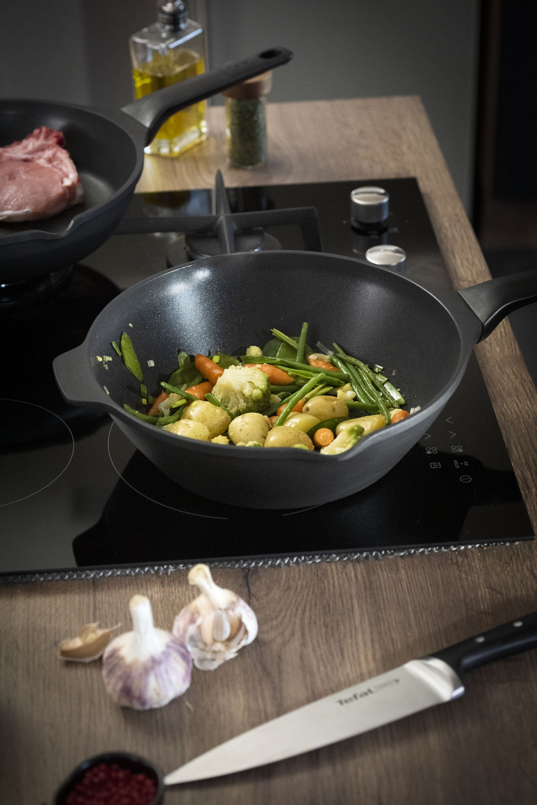 Een wok met groenten staat op een fornuis. Een pan met vlees staat op de achtergrond. Knoflook en een mes liggen op tafel.