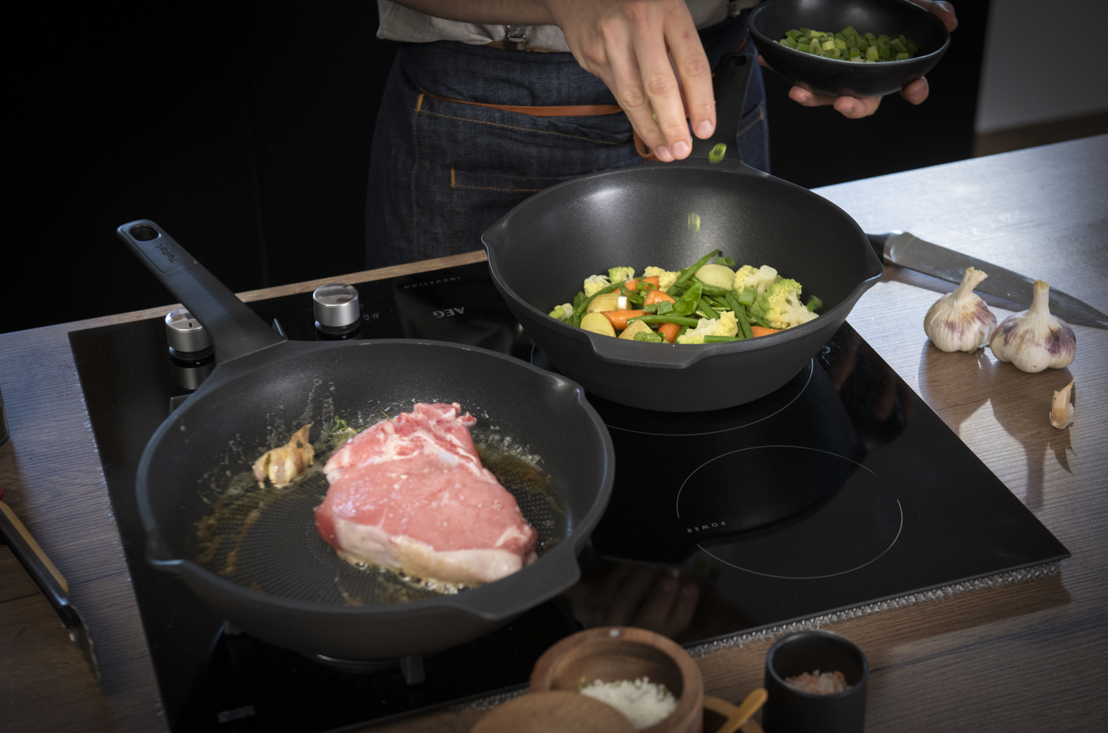 Een persoon kookt. Een biefstuk sist in een pan. Groenten zitten in een wok. Knoflook en kruiden op het aanrecht.