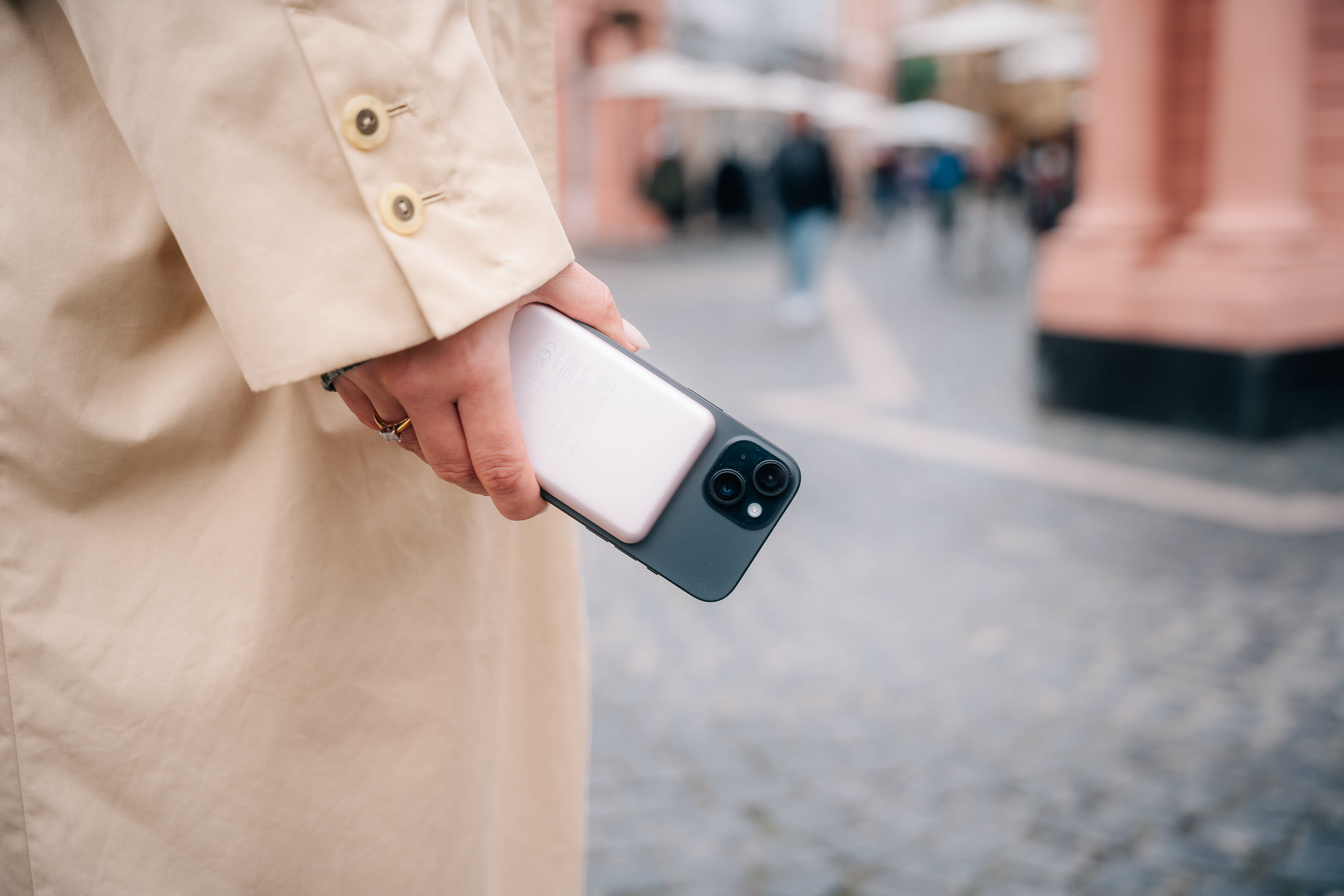 Een persoon houdt een telefoon vast, de achtergrond is wazig met mensen en gebouwen. De persoon draagt een beige jas.