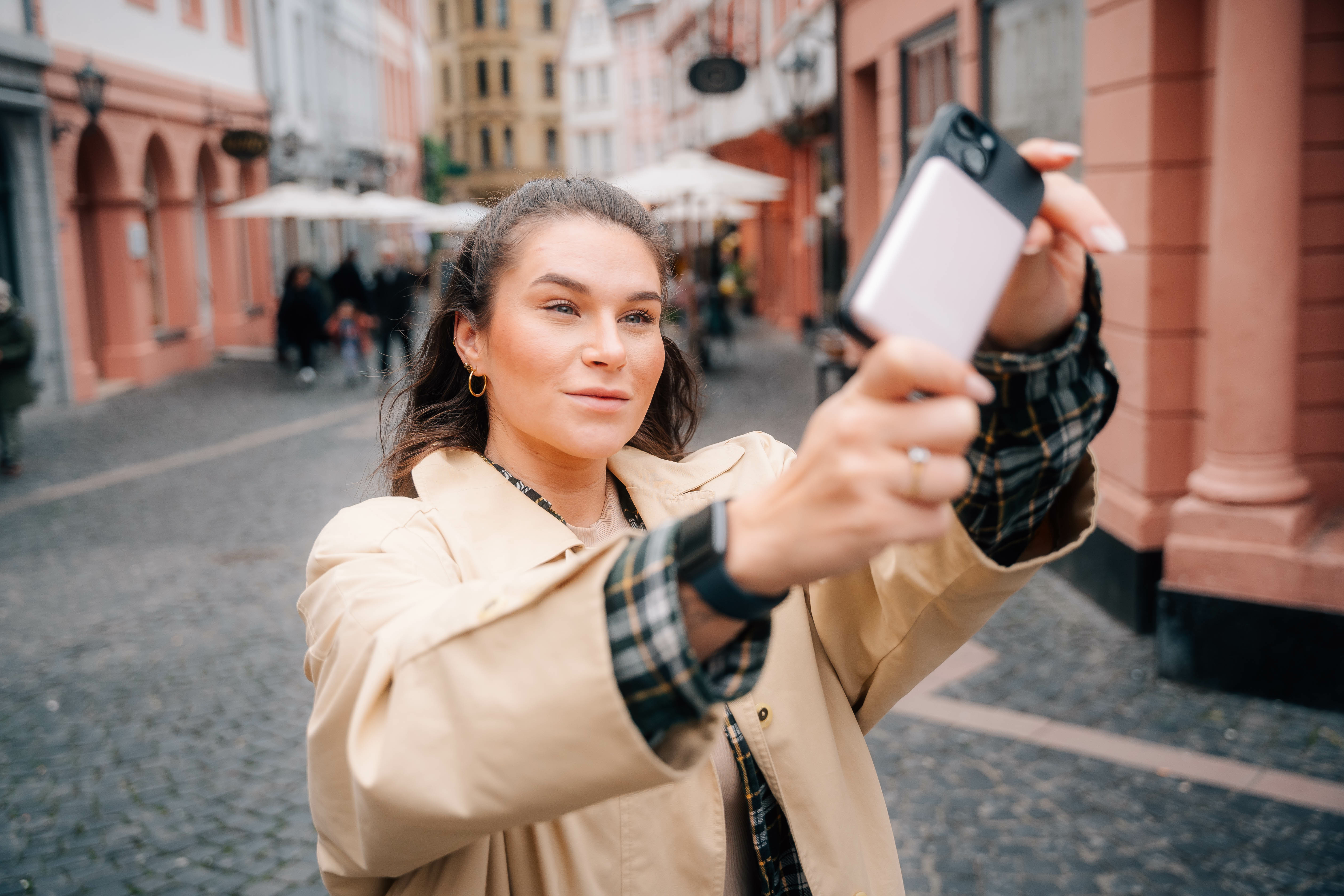 Vrouw maakt selfie met telefoon in straat, beige jas, gouden oorbellen en horloge.