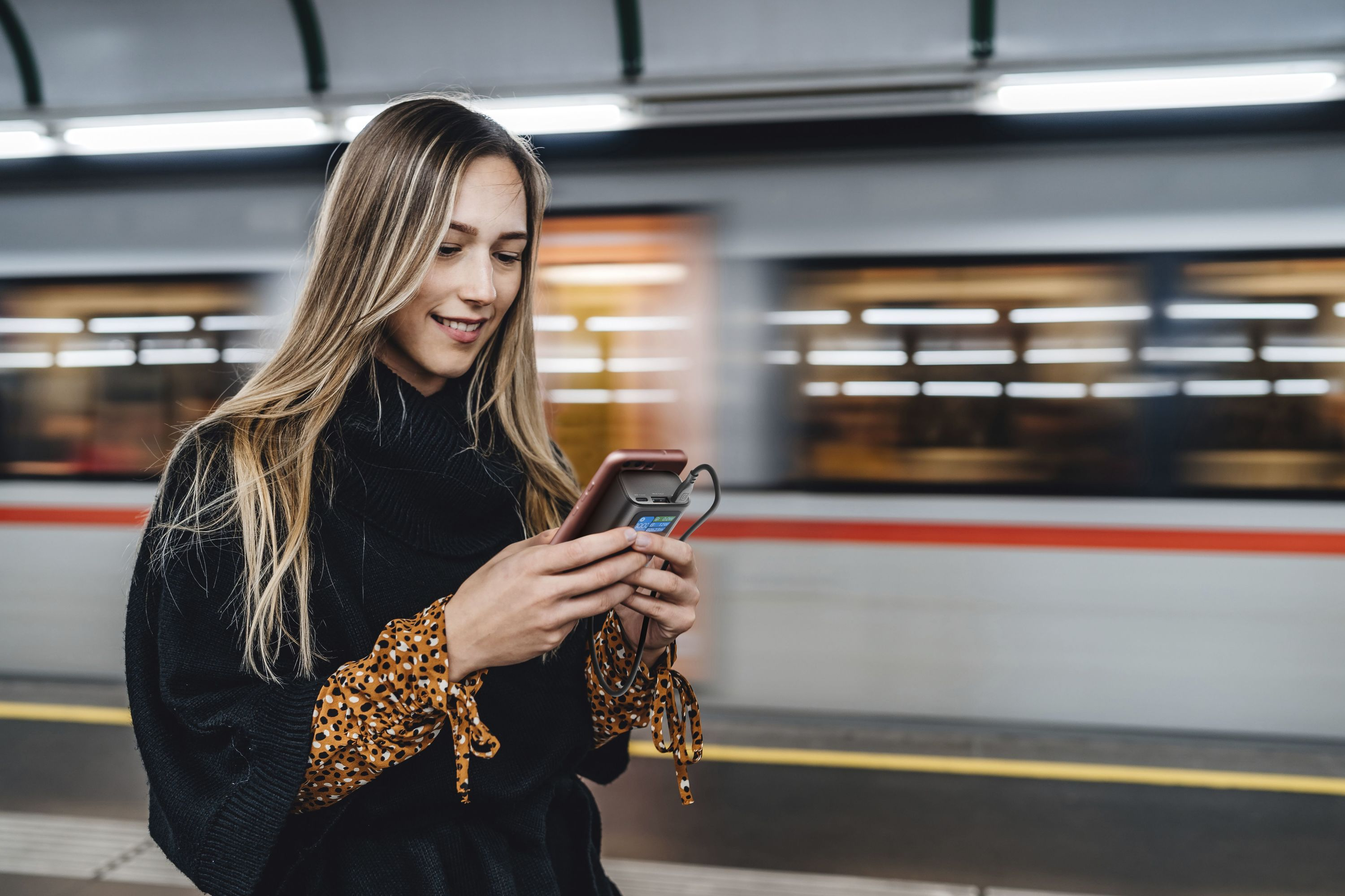 Donna in maglione nero usa il telefono in stazione. Lo sfondo mostra un treno in movimento e dettagli.