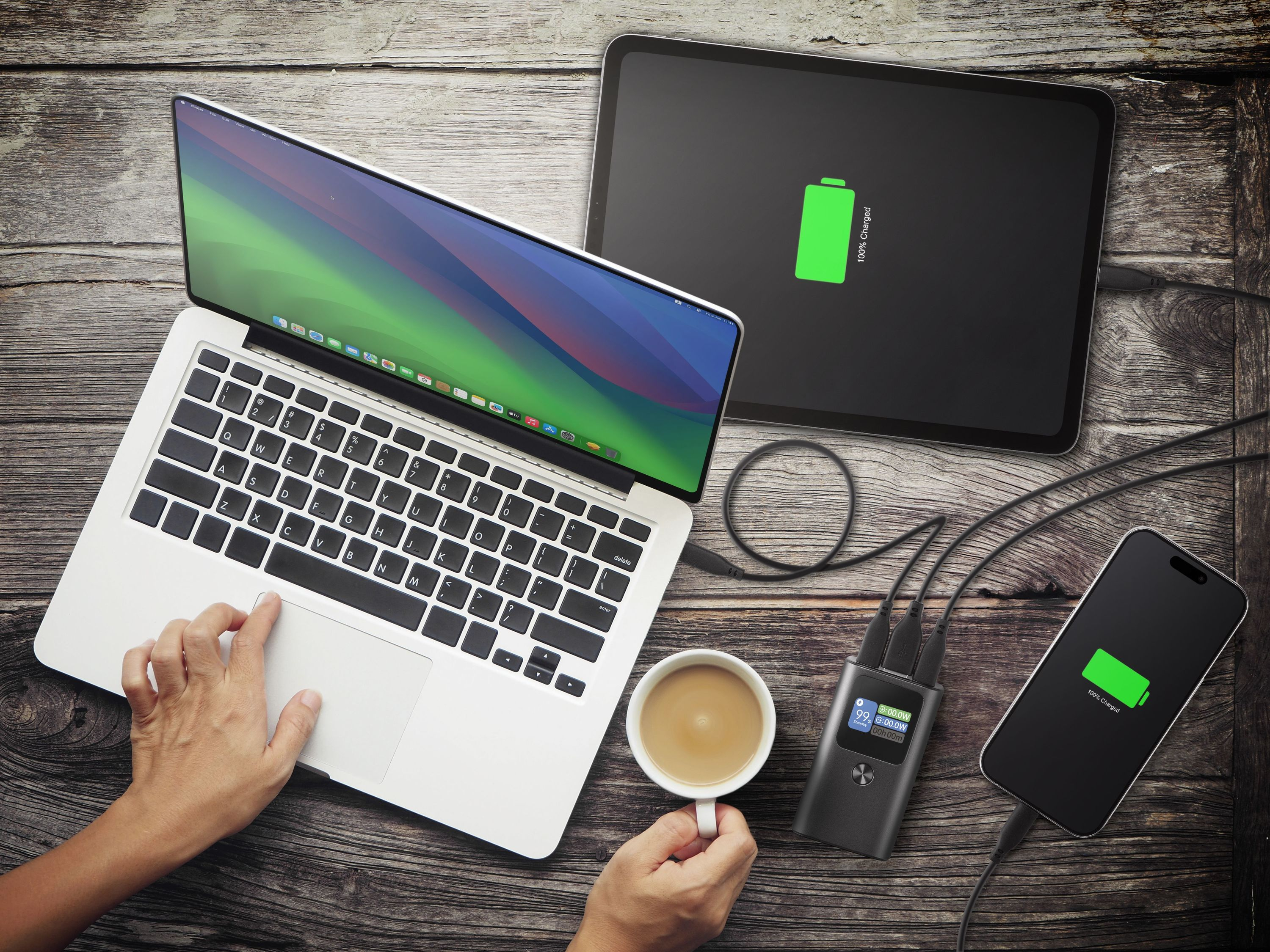 Laptop, tablet e telefono su legno. Tutti carichi. Una mano usa il trackpad. Tazza di caffè.