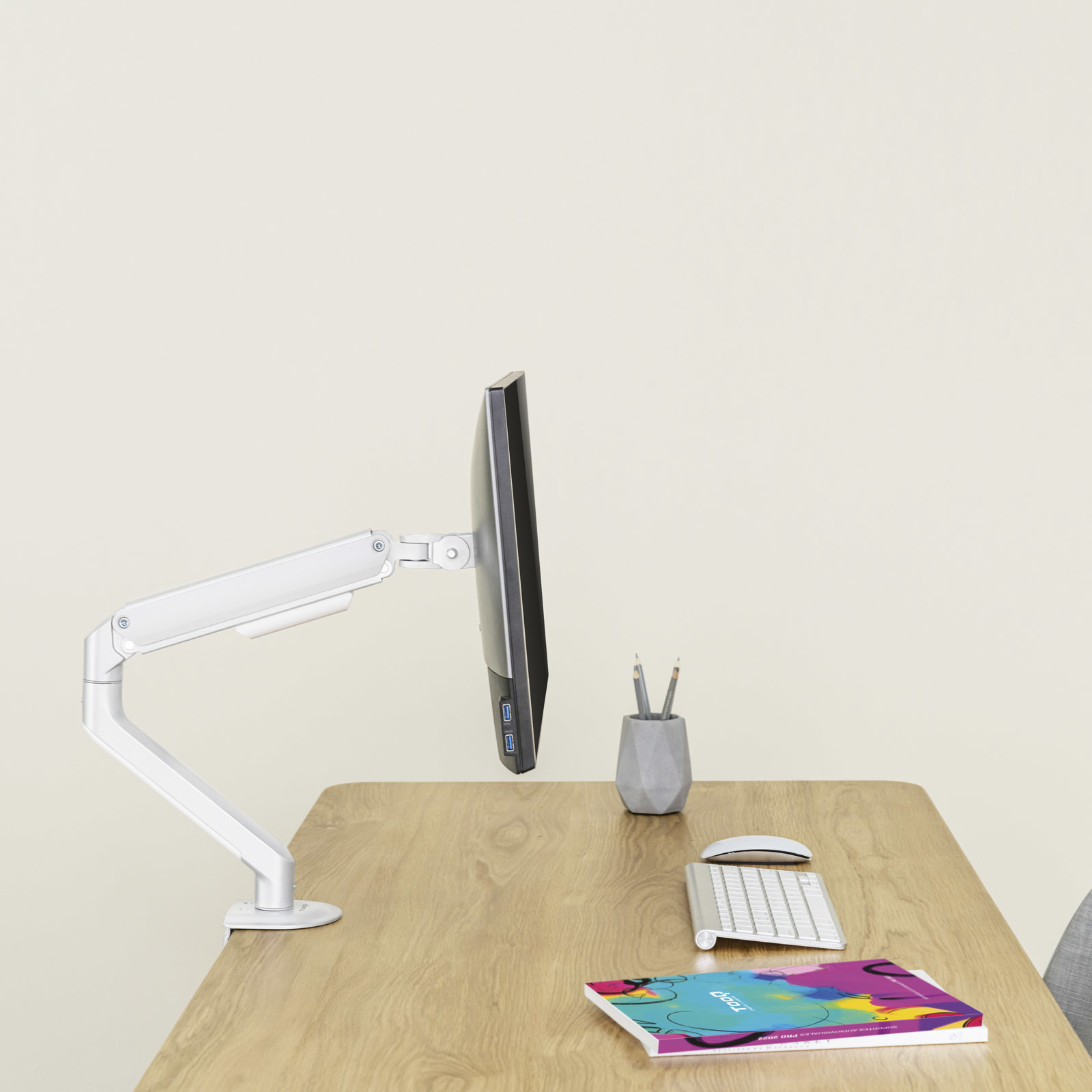Un monitor de computadora está montado en un escritorio con teclado, ratón, portalápices y un libro.