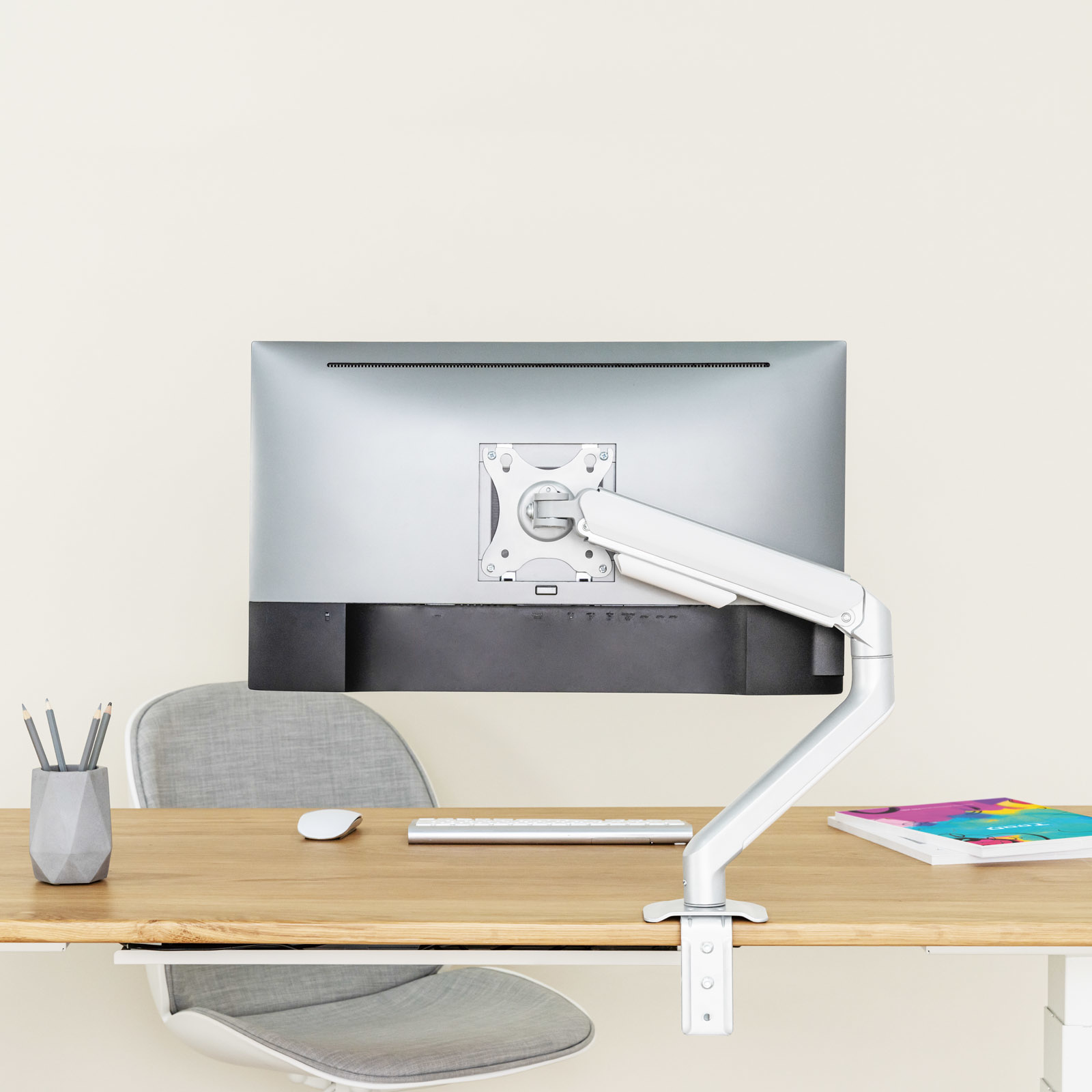 Un monitor de ordenador con soporte, un escritorio con teclado, ratón, lápices y un libro sobre un fondo claro.