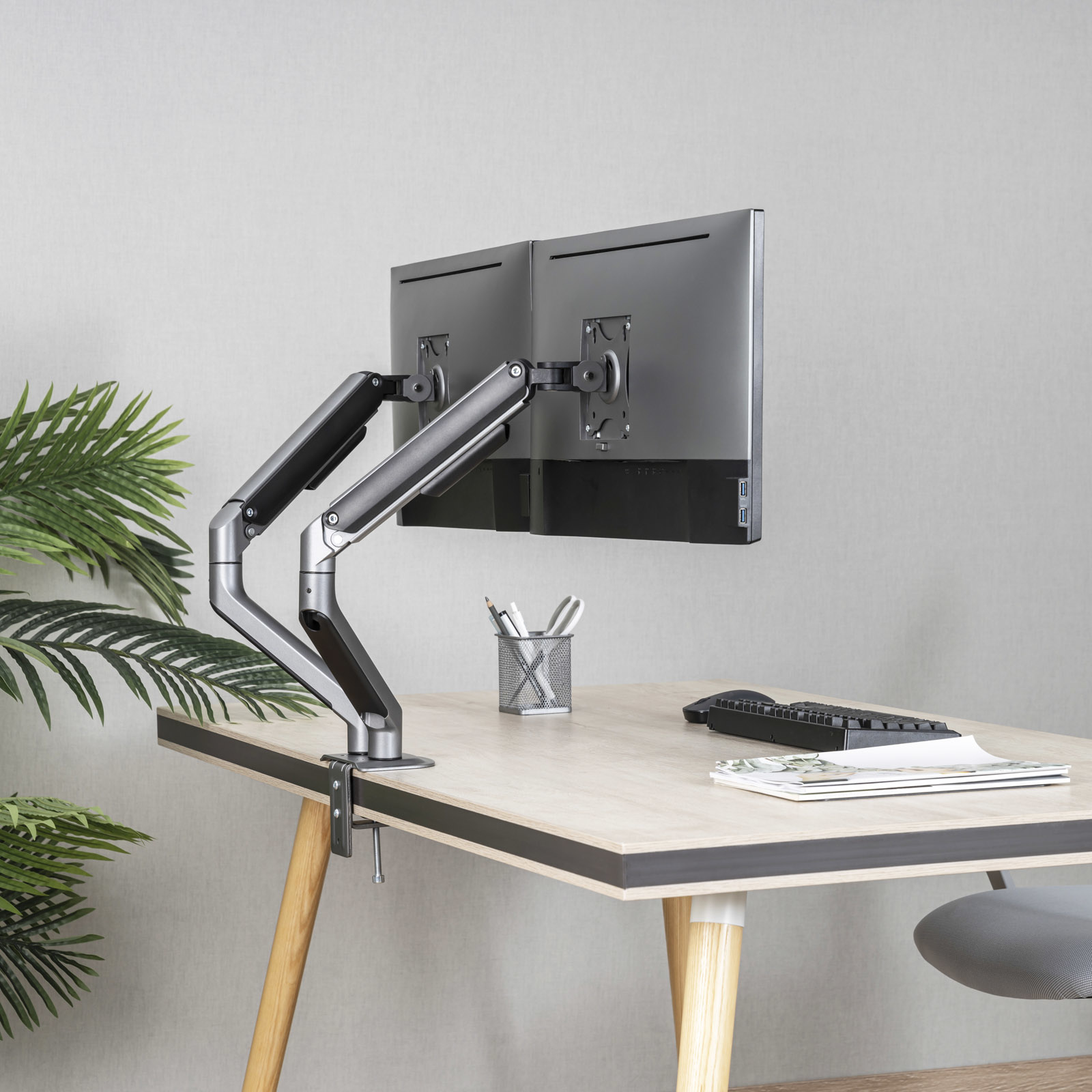 Escritorio con dos monitores, brazos de monitor, teclado y un portalápices. También hay una planta y una silla gris.