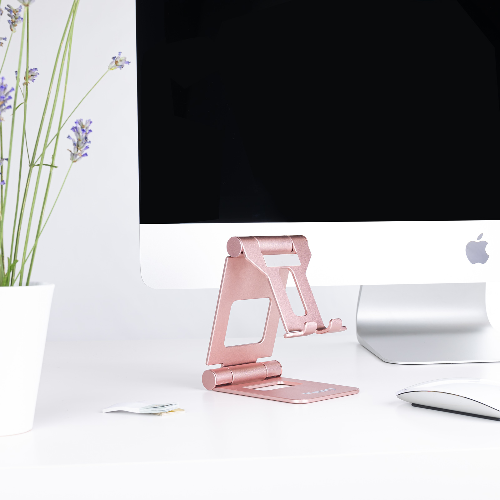 Un soporte para teléfono oro rosa, computadora, lavanda y ratón sobre un escritorio blanco.