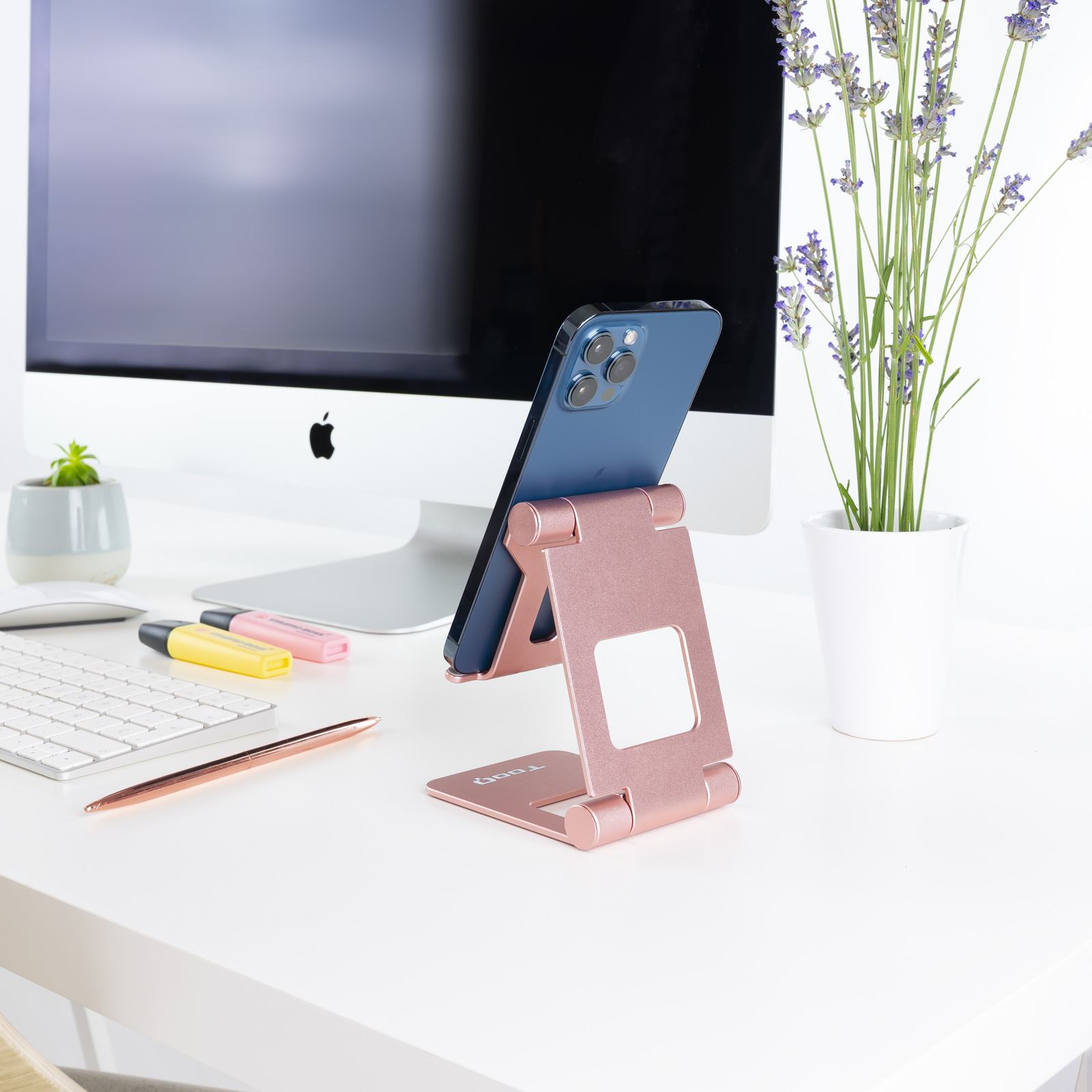 Teléfono azul en soporte rosa. Monitor Apple, teclado, bolígrafo y flores en escritorio blanco.