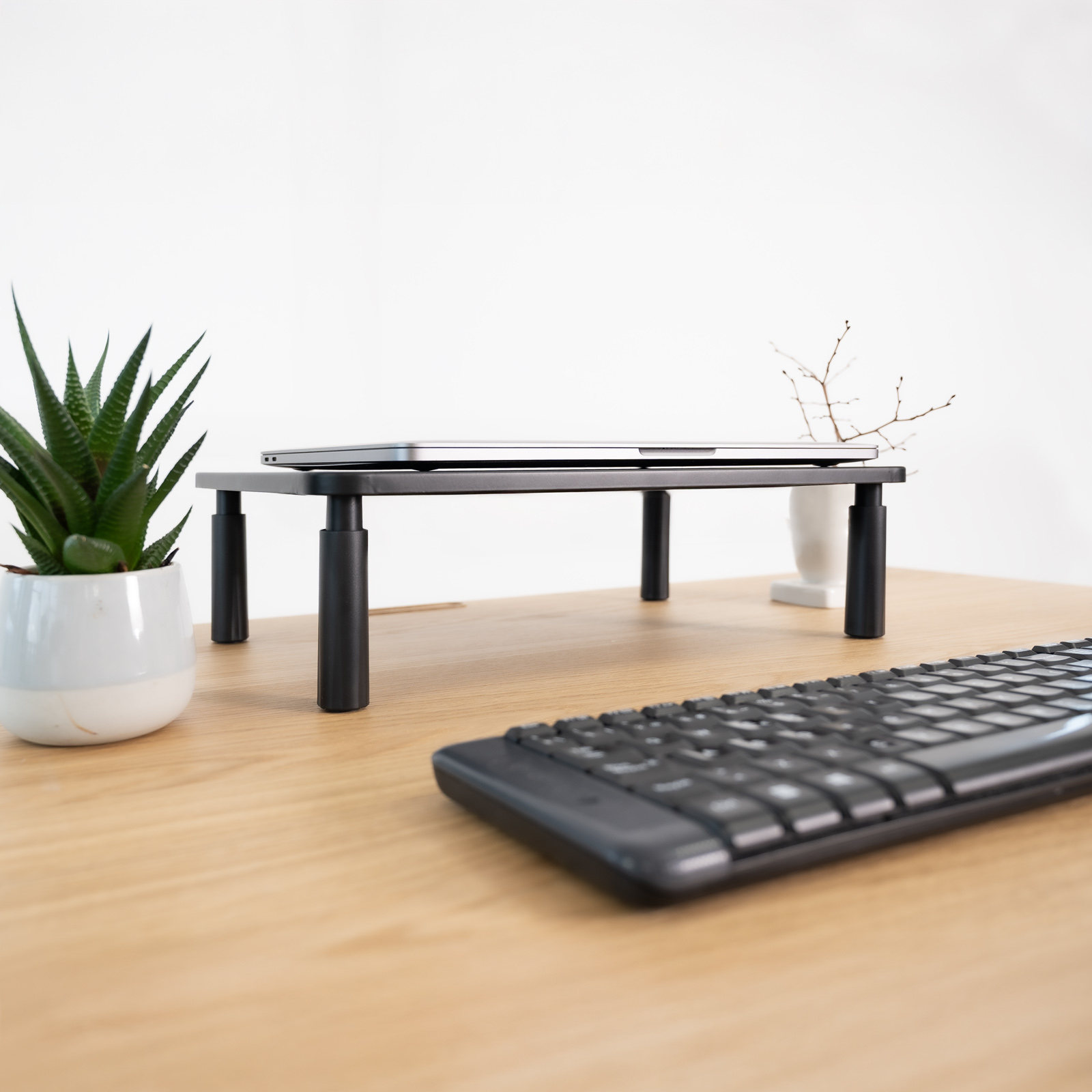 Un laptop è su un supporto nero su una scrivania di legno. Sulla scrivania ci sono anche una tastiera e una pianta.