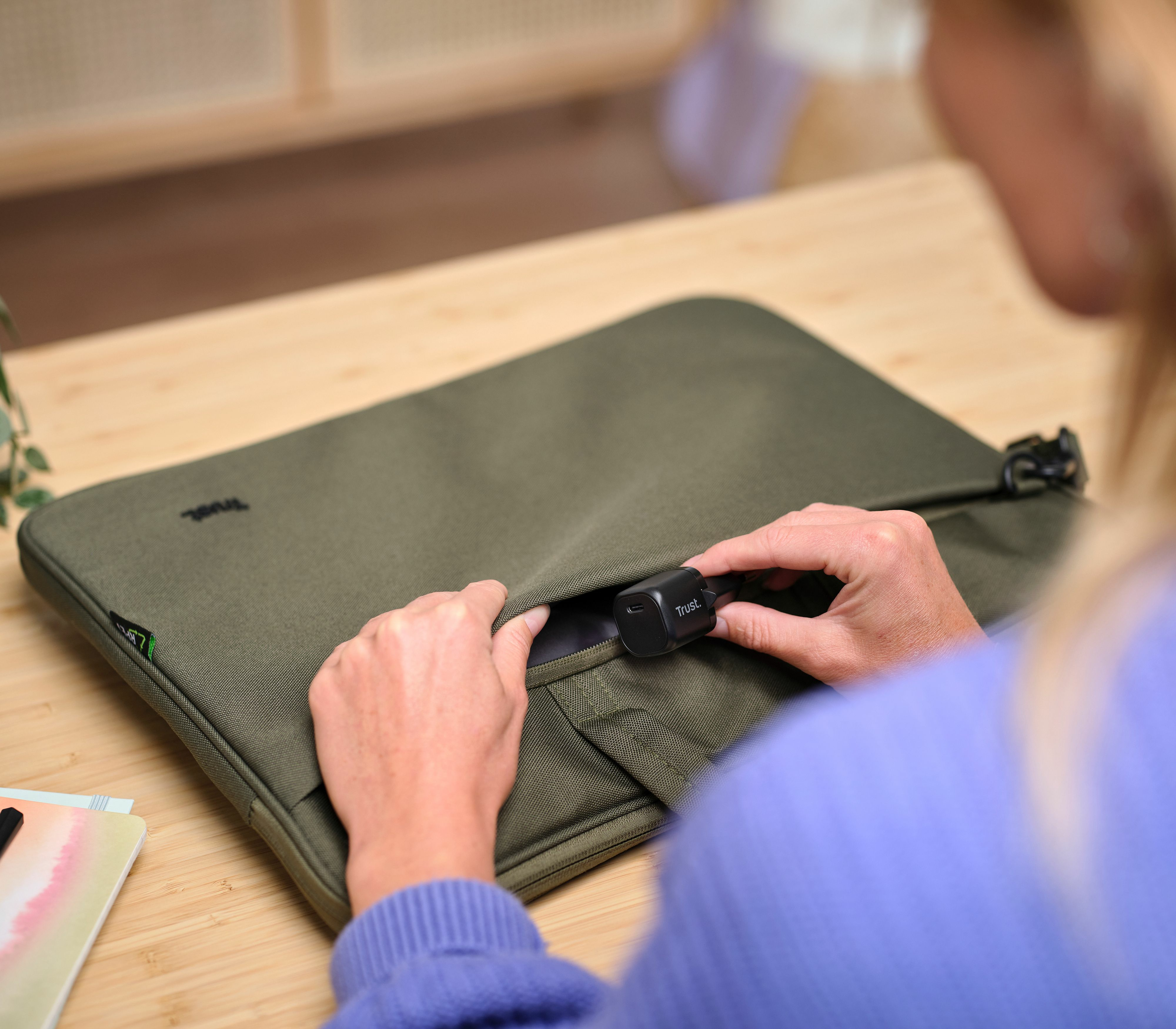 Una persona sostiene un dispositivo Trust negro y lo coloca en una funda verde para portátil sobre una mesa de madera.