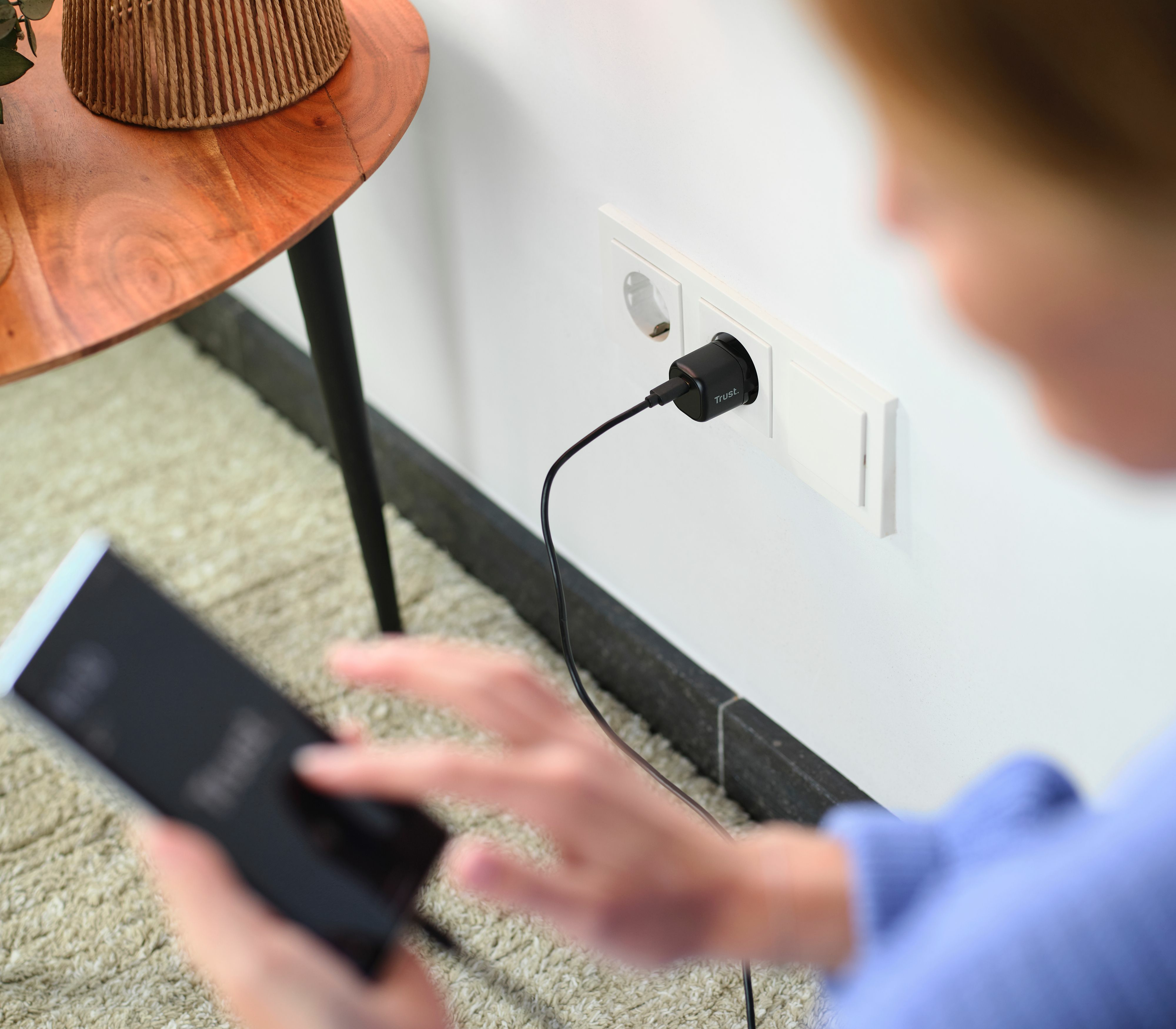 Un teléfono se está cargando. Un cargador Trust está enchufado a la pared. Una persona sostiene el teléfono.