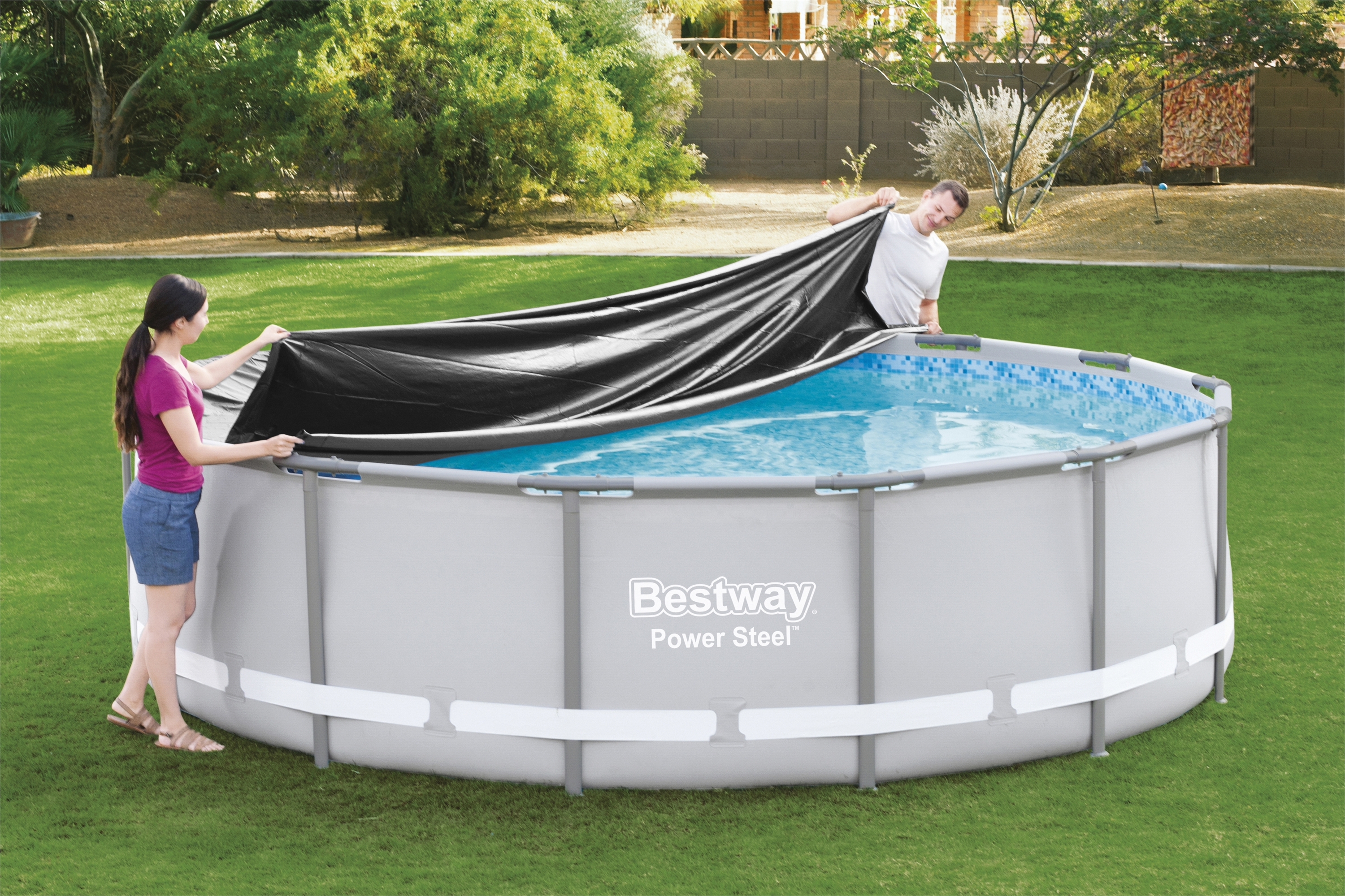Zwei Personen bedecken einen runden Pool mit einer schwarzen Abdeckung. Grünes Gras umgibt den Pool. Bäume sind im Hintergrund.