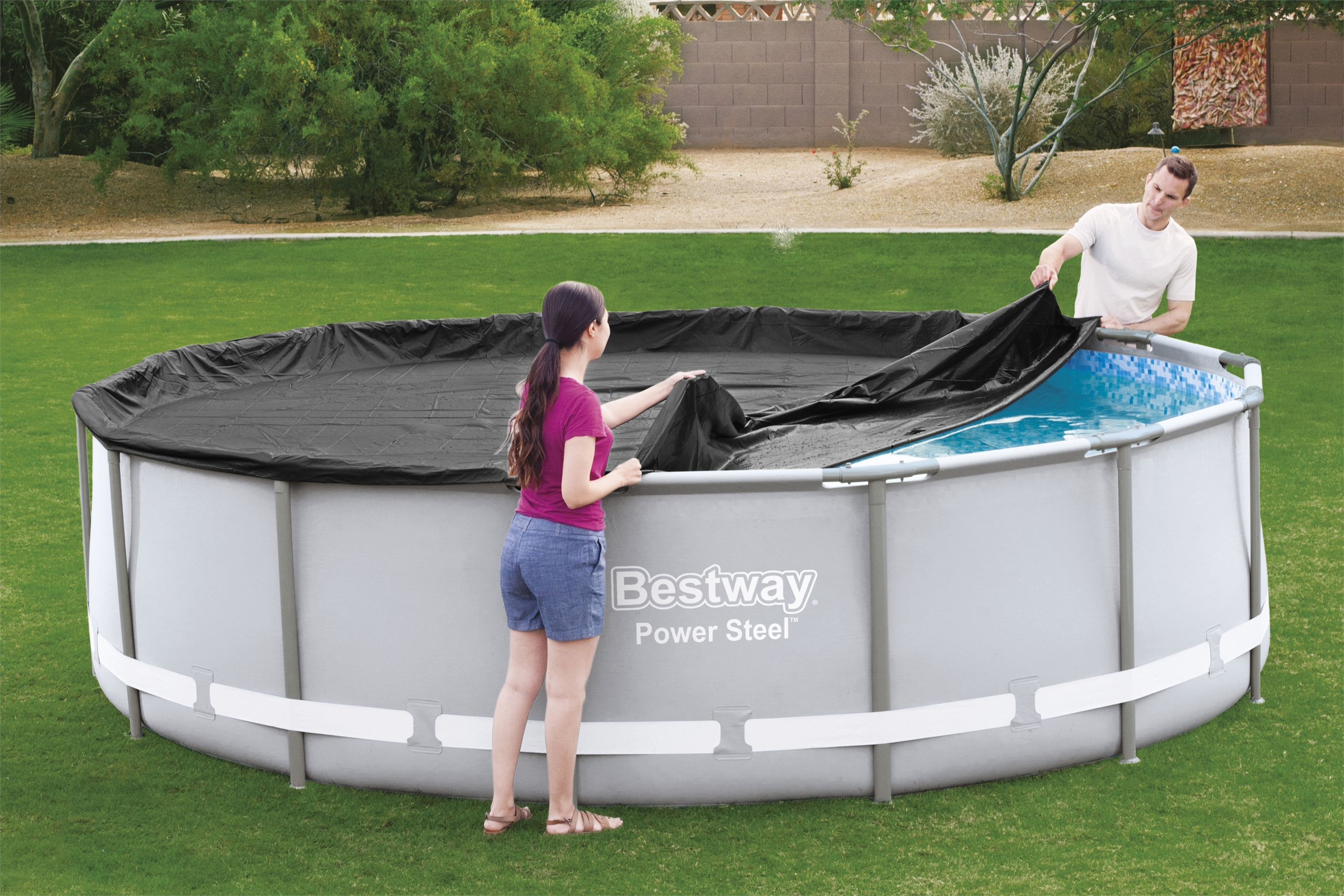 Zwei Personen entfernen eine schwarze Poolabdeckung von einem runden, oberirdischen Pool auf Rasen.