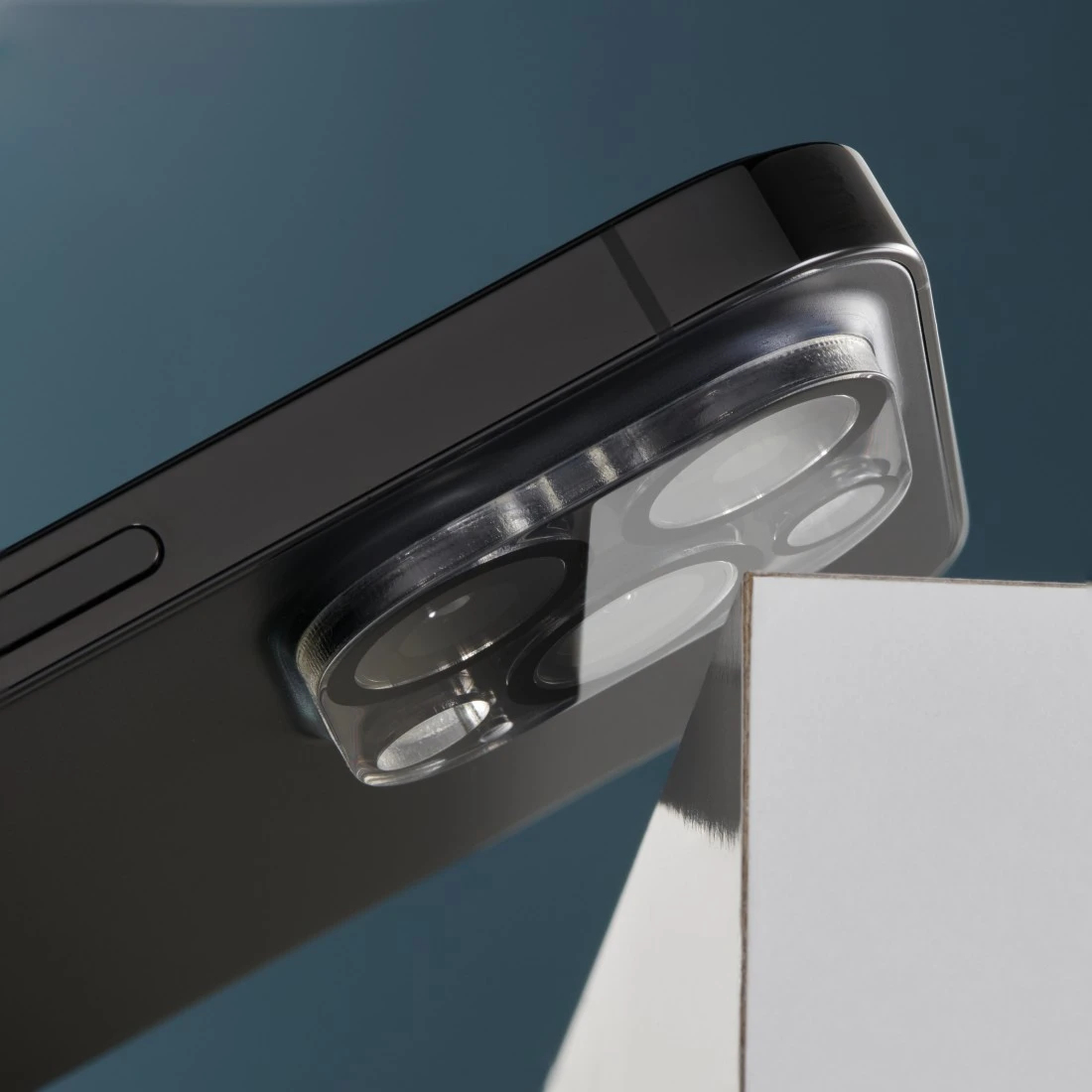 Primo piano di uno smartphone nero con protezione trasparente per lenti. Il telefono poggia su una superficie bianca su sfondo blu.