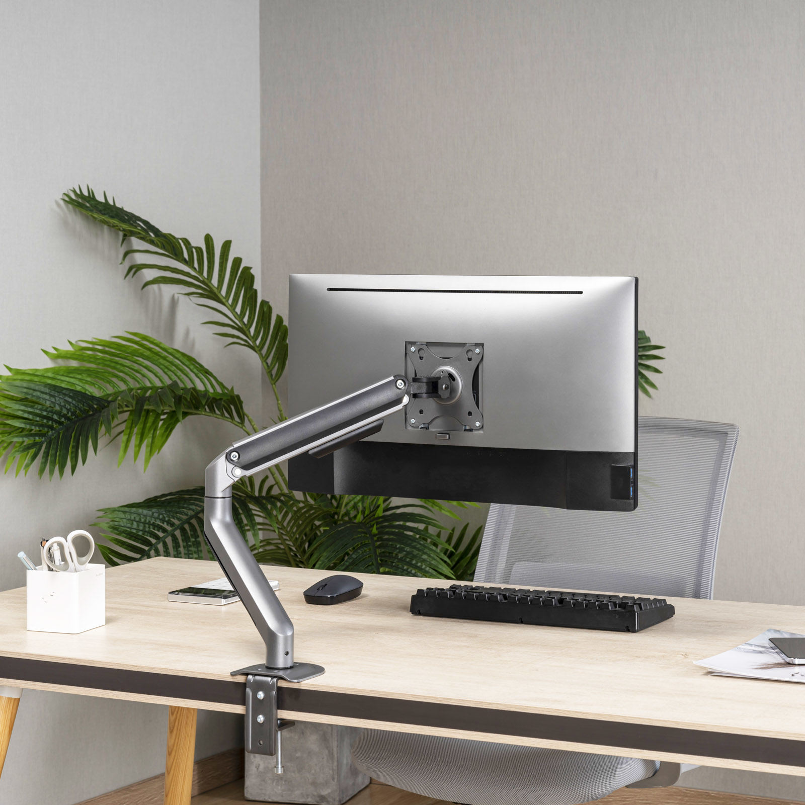 Un monitor de computadora está montado en un escritorio. Teclado, mouse y otros suministros de oficina están en el escritorio.