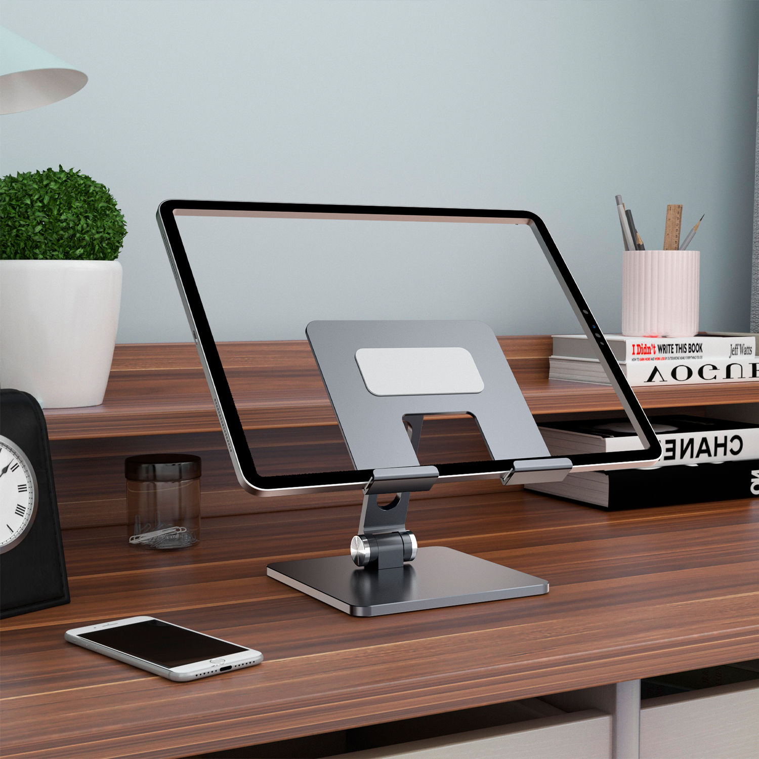 Tablet auf Ständer, Schreibtisch mit Telefon, Uhr, Büchern und Pflanze. Grauer Tablet-Ständer. Holzschreibtisch mit Schubladen und Wand im Hintergrund.