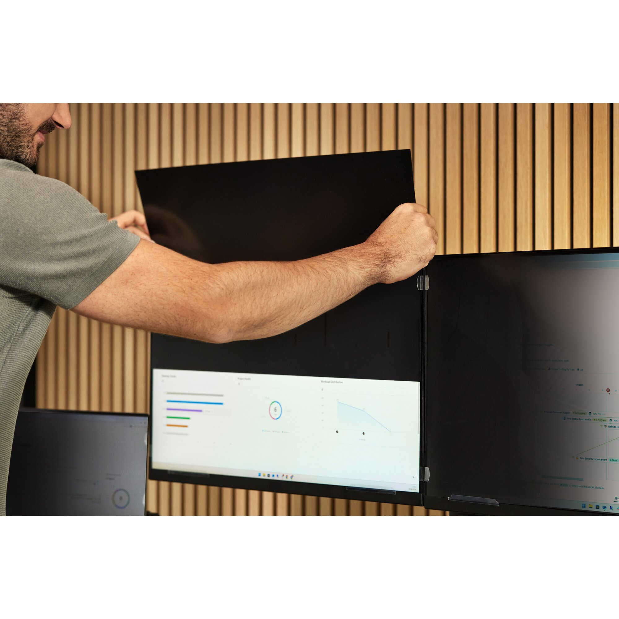 Un hombre coloca una pantalla de privacidad negra en un monitor. El fondo es una pared de madera.