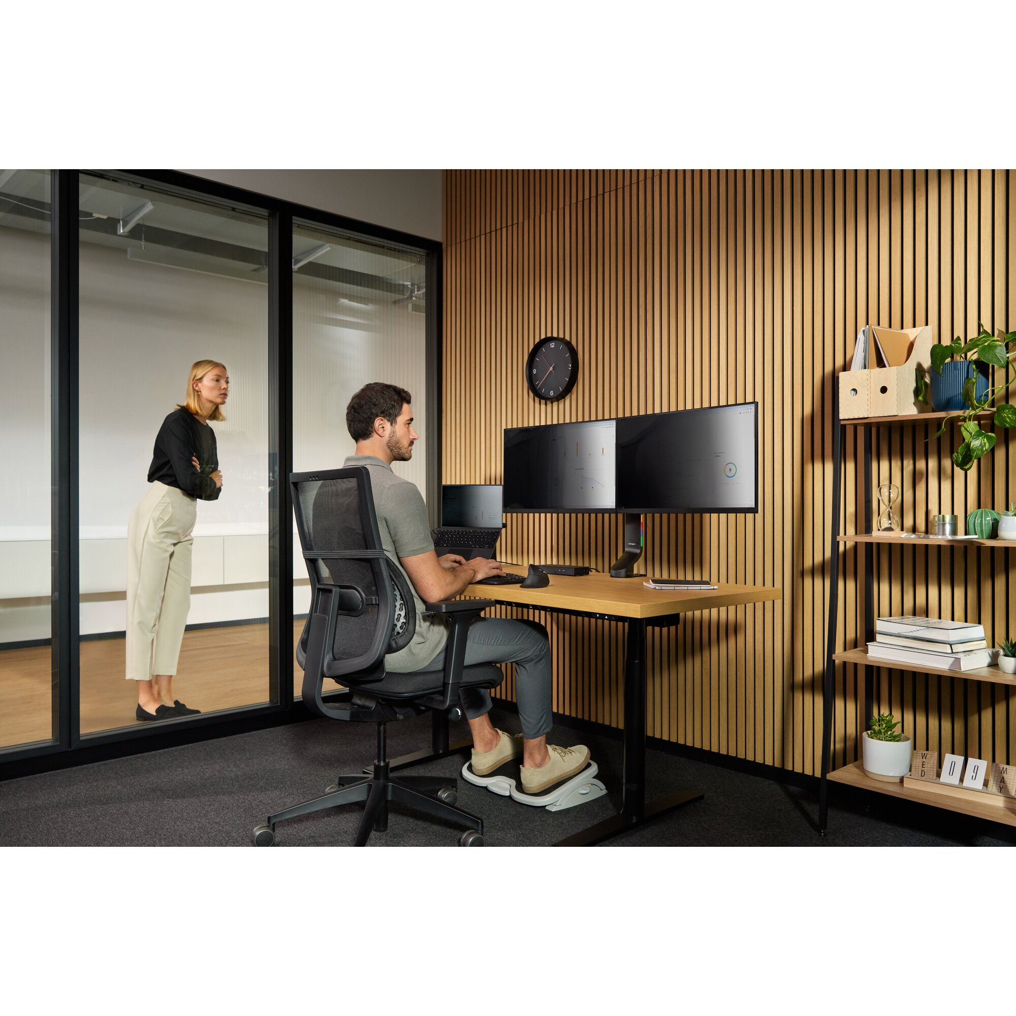 Mann arbeitet an einem Schreibtisch mit zwei Monitoren. Frau beobachtet durch Glas. Holzwand, Uhr und Regale mit Pflanzen im Hintergrund.