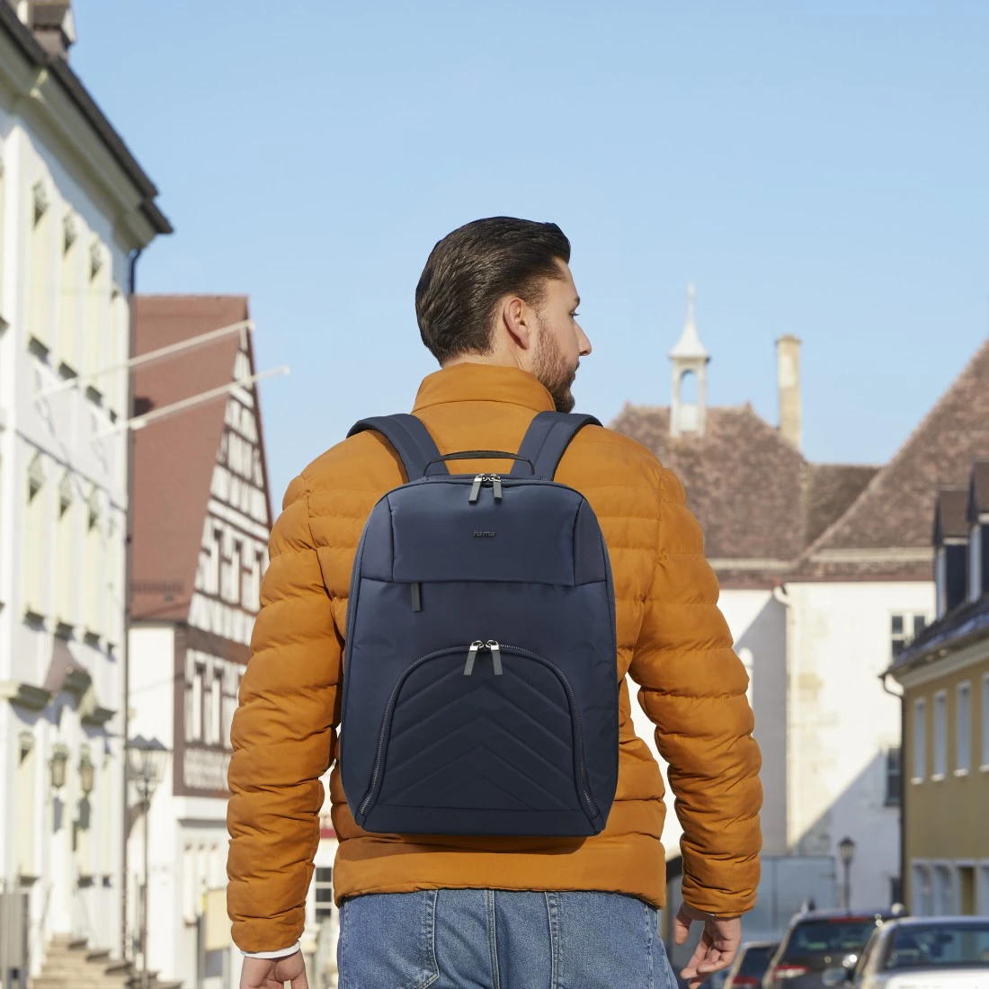 Un hombre con chaqueta naranja y mochila azul camina por una calle. Los edificios bordean la calle, el cielo está despejado y azul.