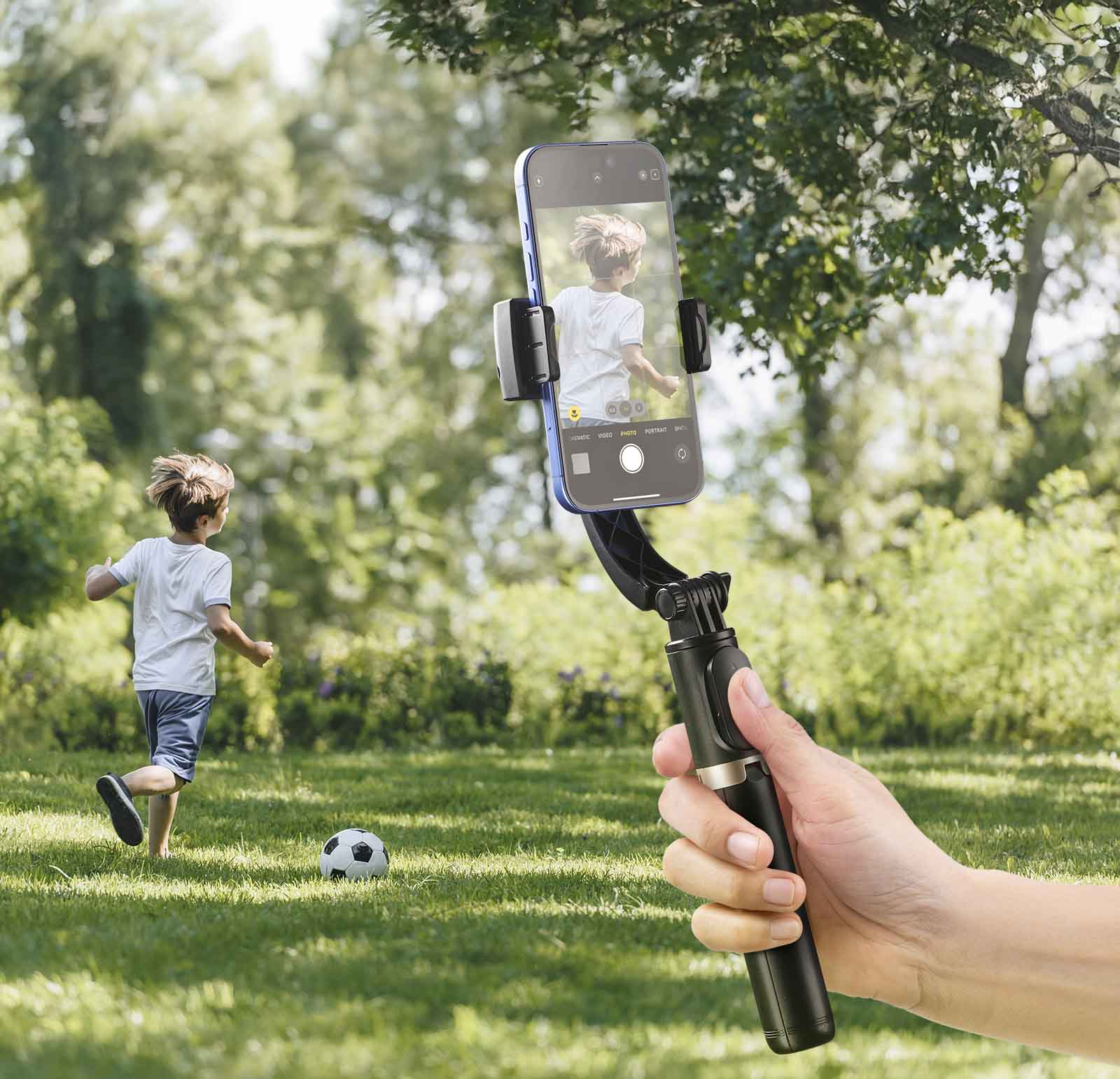 Eine Person hält ein Telefon an einem Selfie-Stick. Ein Junge rennt auf einen Fußball auf dem Rasen zu. Grüne Bäume im Hintergrund.
