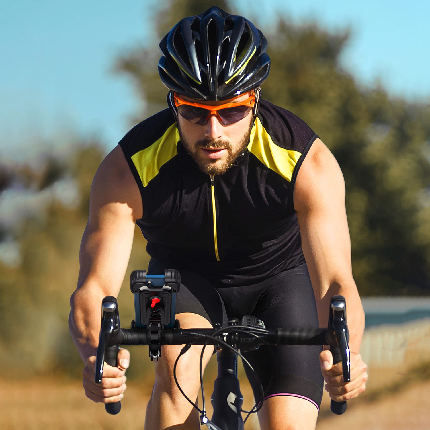 Kolarz na rowerze szosowym ma na sobie kask i okulary przeciwsłoneczne. Telefon jest zamontowany na kierownicy. Tło to rozmyte drzewa i niebo.