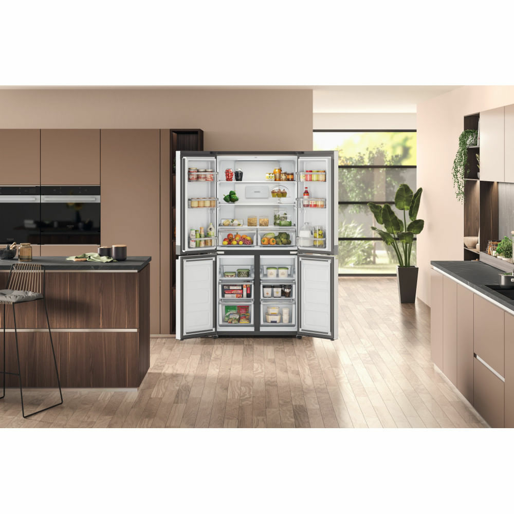 Frigorifero aperto in una cucina moderna, pieno di cibo. Pavimento in legno, armadi, isola e una pianta alta sullo sfondo.
