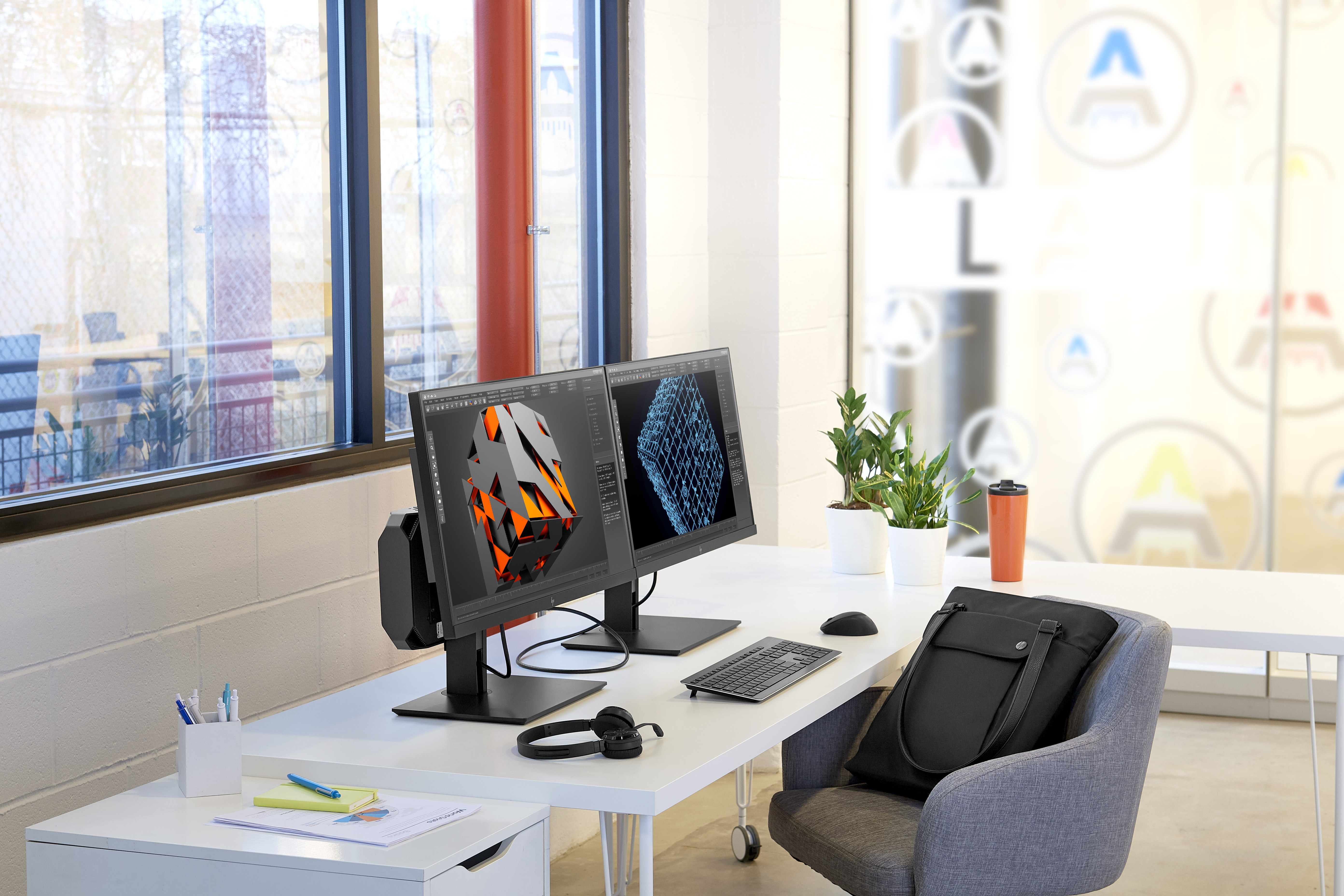 Scrivania con due monitor che mostrano modelli 3D. Una borsa, delle piante e una tazza da caffè sulla scrivania.