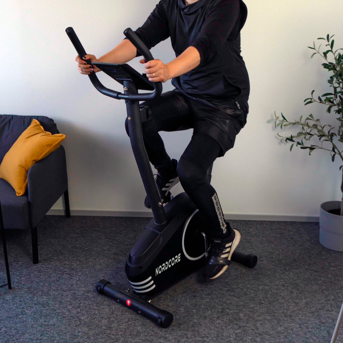 Osoba jedzie na czarnym rowerze treningowym. Ma na sobie czarny strój sportowy. Żółta poduszka leży na szarej sofie.
