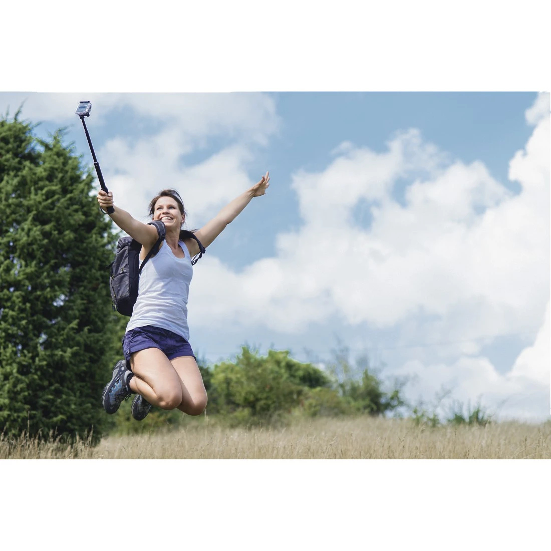 Frau springt, hält Selfie-Stick, Rucksack. Trägt Shorts, Tanktop und Schuhe. Blauer Himmel mit Wolken, Feld und Bäume im Hintergrund.