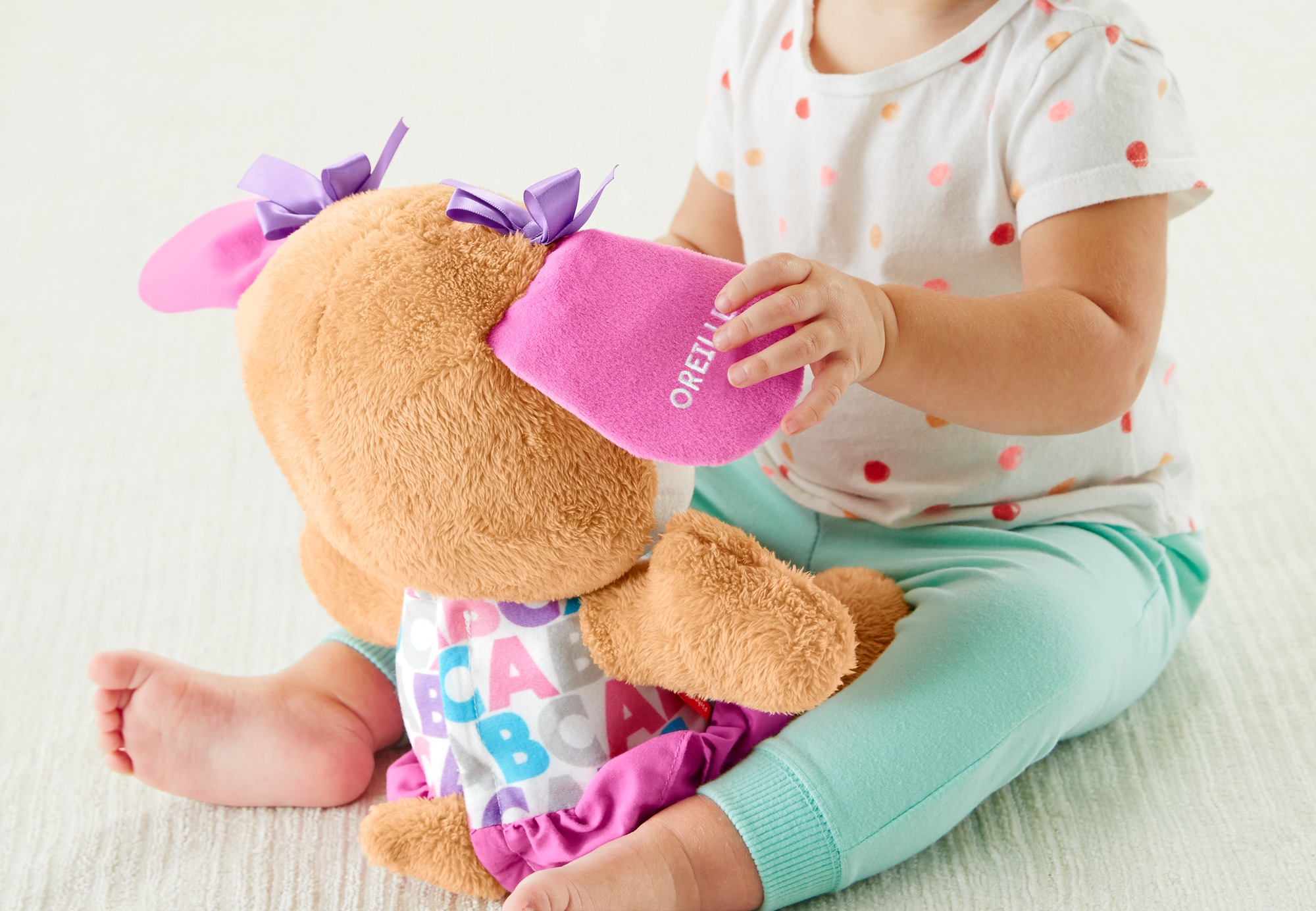 Un bebé sostiene un peluche con lazos morados y orejas rosas. El juguete es marrón. El bebé viste una camisa de lunares y pantalones menta.