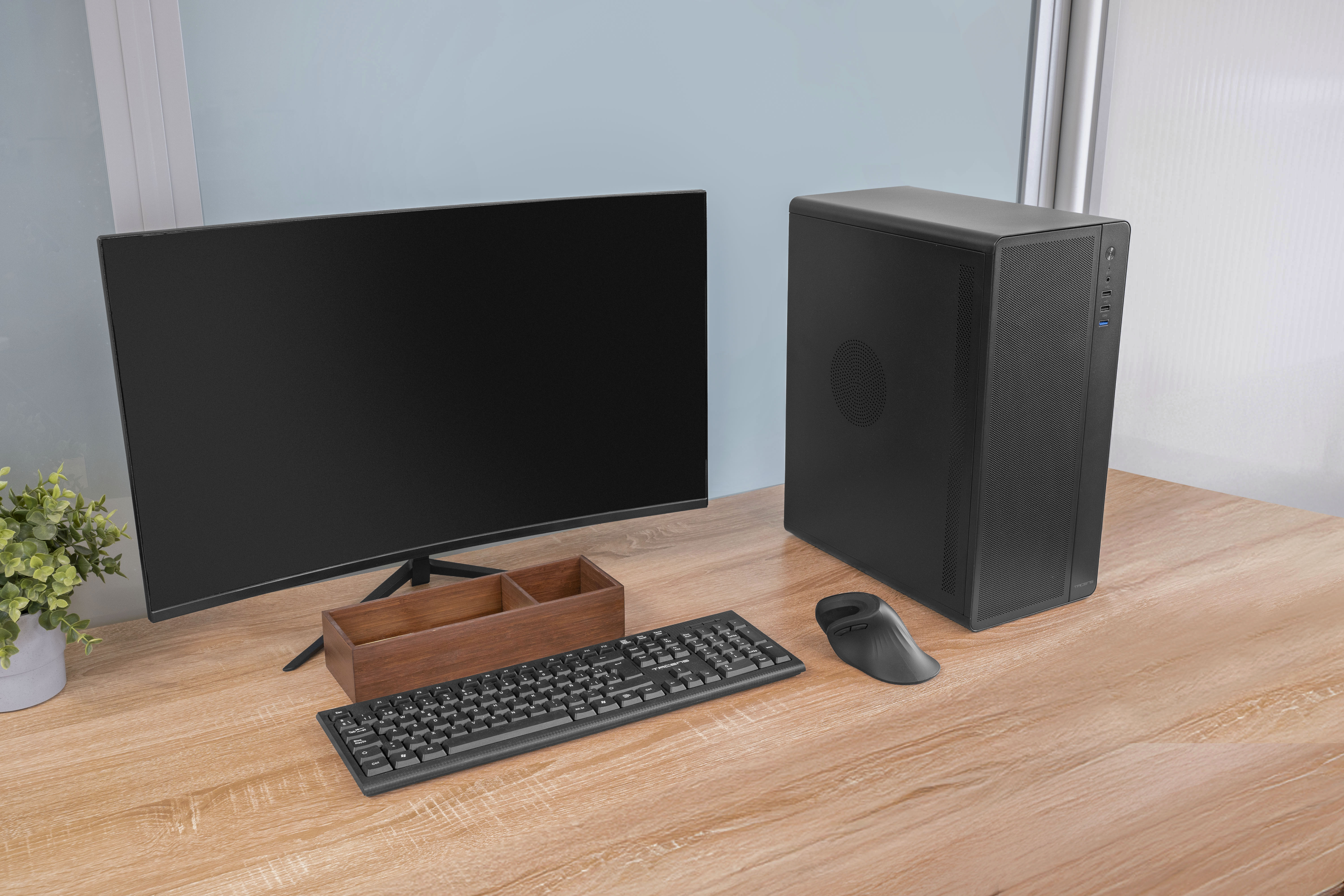 Una configuración de computadora en un escritorio de madera. Incluye monitor, caja de PC, teclado, ratón y planta.