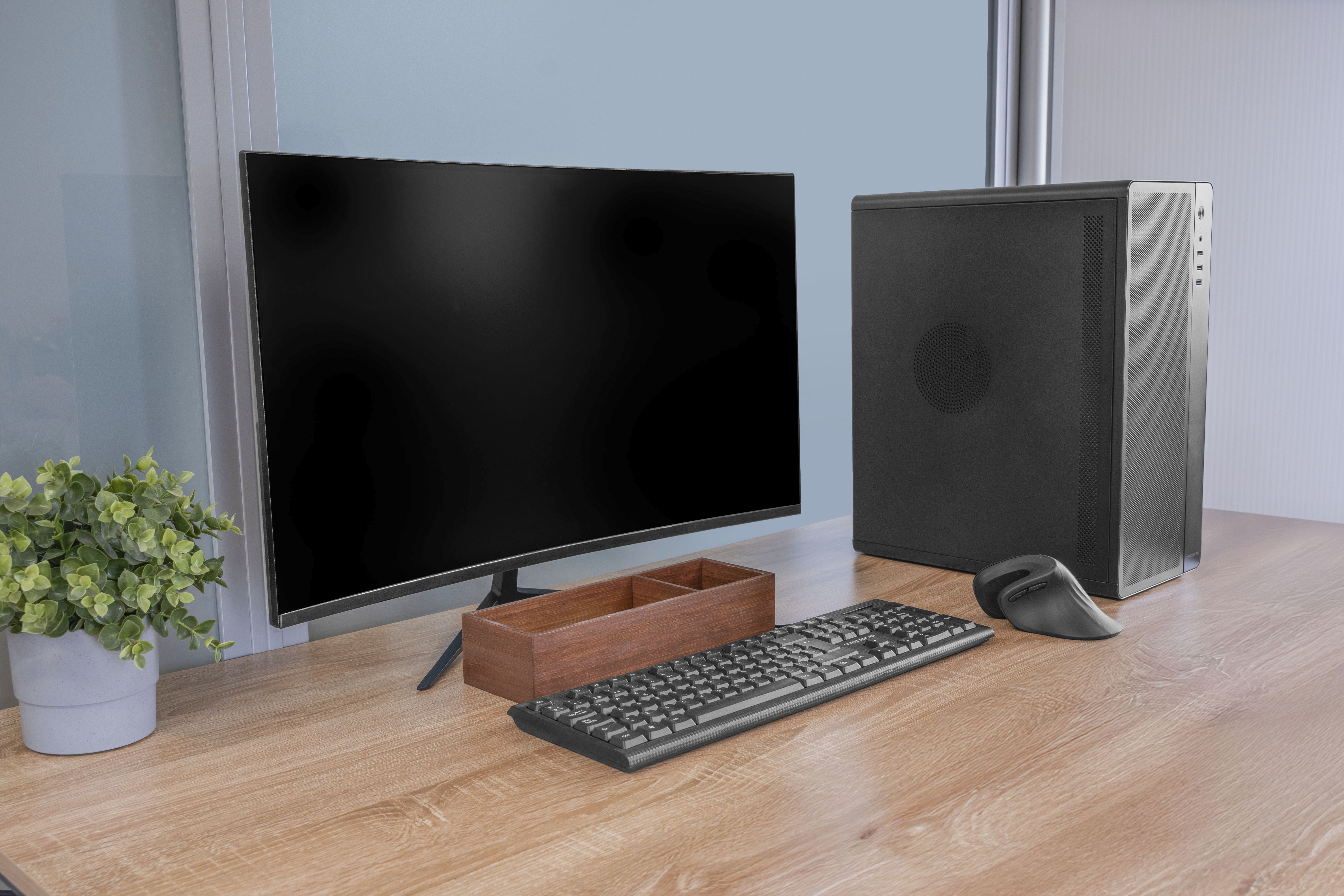 Una configuración de computadora con monitor, carcasa de PC, teclado, ratón y planta en un escritorio de madera.