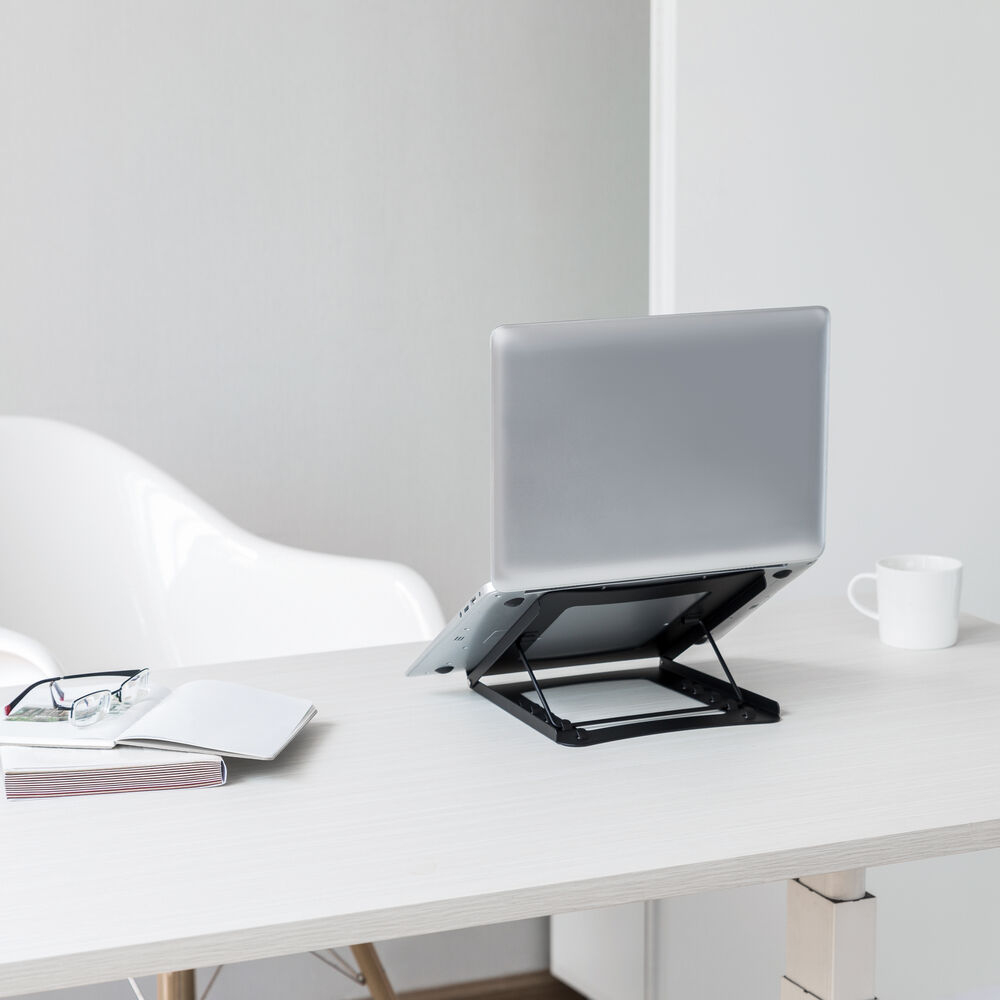 A laptop sits on a stand on a white desk. Beside it are glasses, a notebook, and a mug. A white chair is in the background.