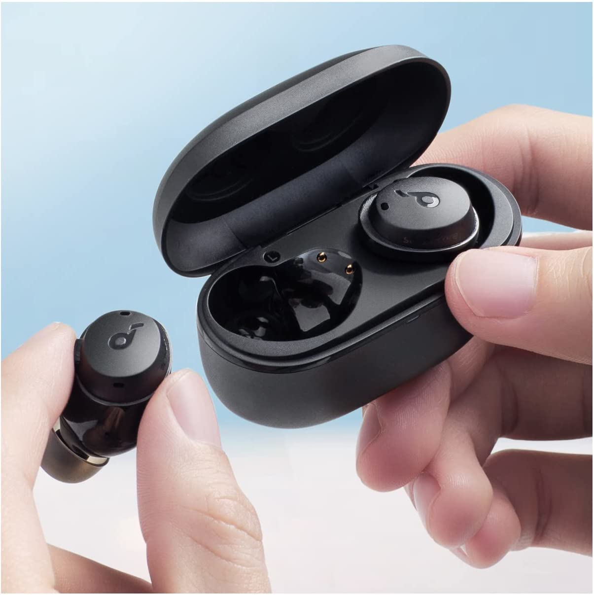 Manos sosteniendo auriculares inalámbricos negros y un estuche de carga. Un auricular se sostiene por separado. El fondo es azul claro.