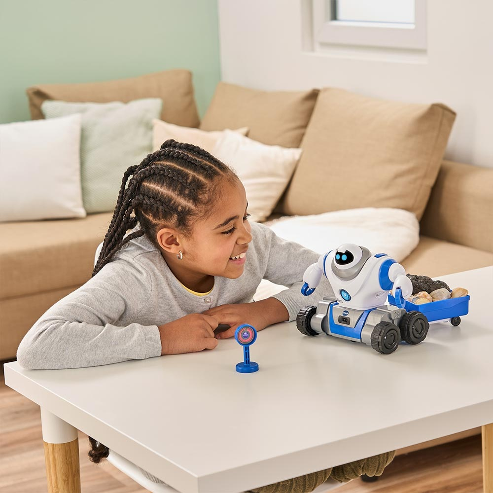 Une fille sourit à un robot sur une table. Le robot a une charrette bleue avec des roches. Une petite cible est également sur la table.