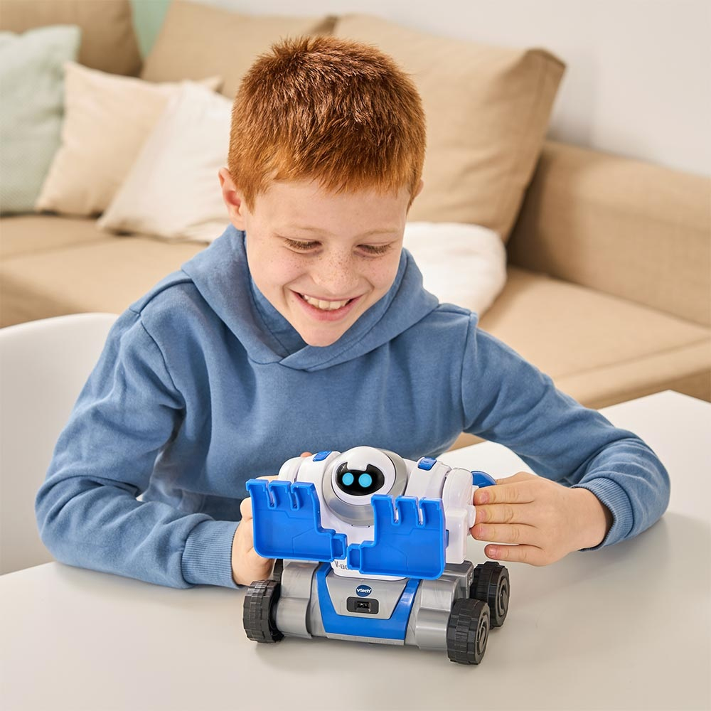 Un garçon souriant aux cheveux roux joue avec un jouet robot blanc et bleu sur une table blanche. Un canapé est en arrière-plan.
