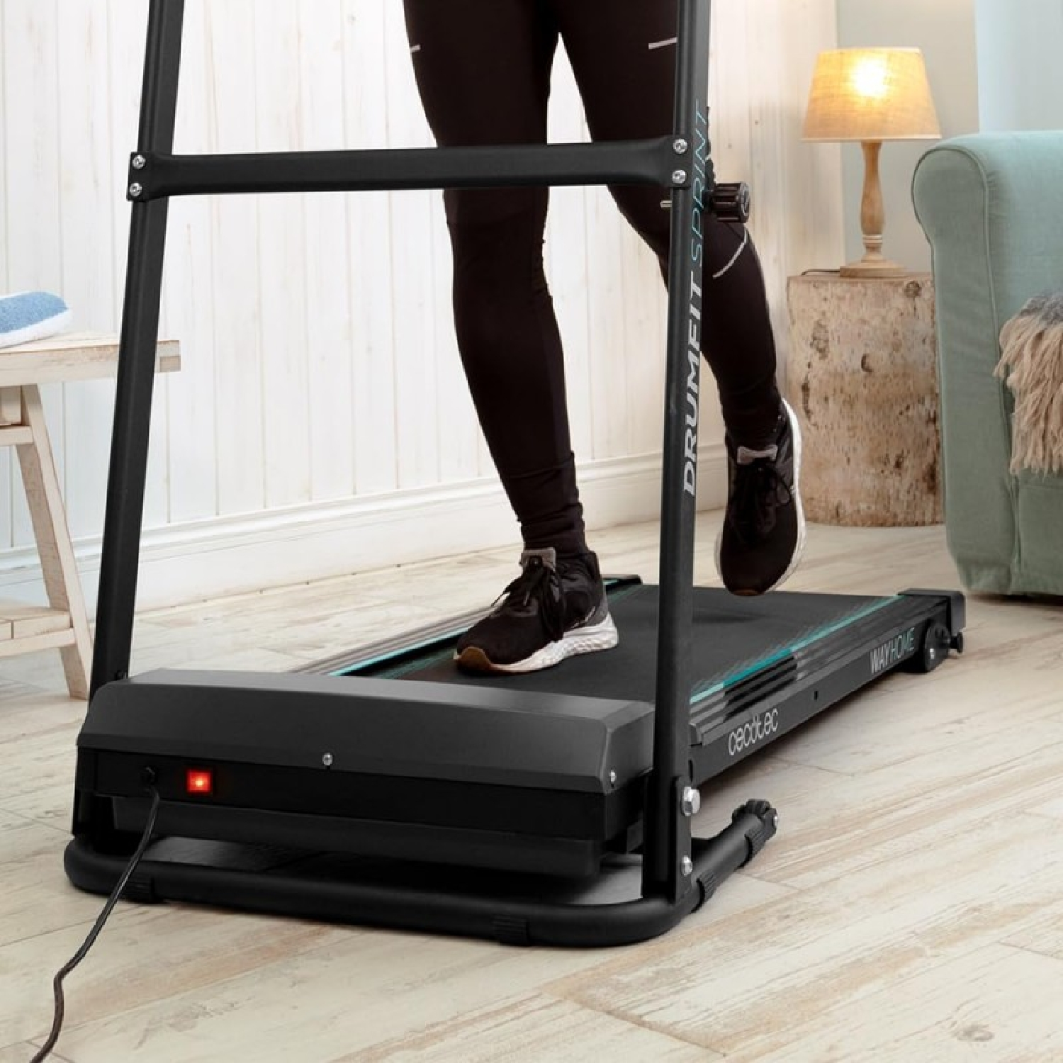 Una persona corre su un tapis roulant nero. La persona indossa leggings e scarpe nere. Una lampada e un divano sono sullo sfondo.