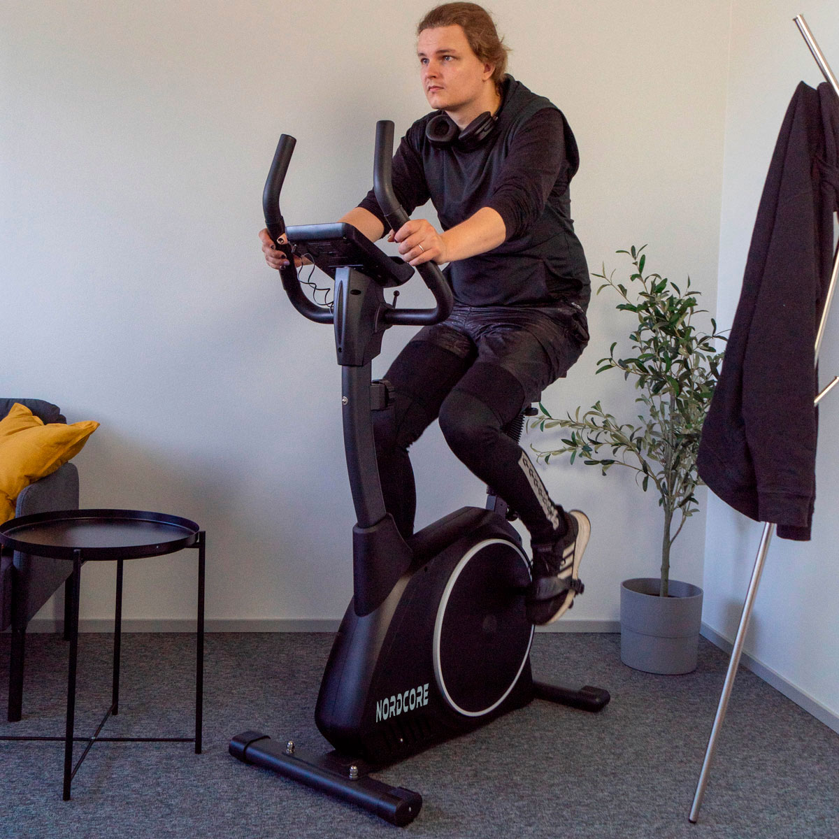 Mężczyzna w czarnym stroju sportowym jeździ na rowerze stacjonarnym. Rower jest w pokoju z sofą, stolikiem i rośliną.
