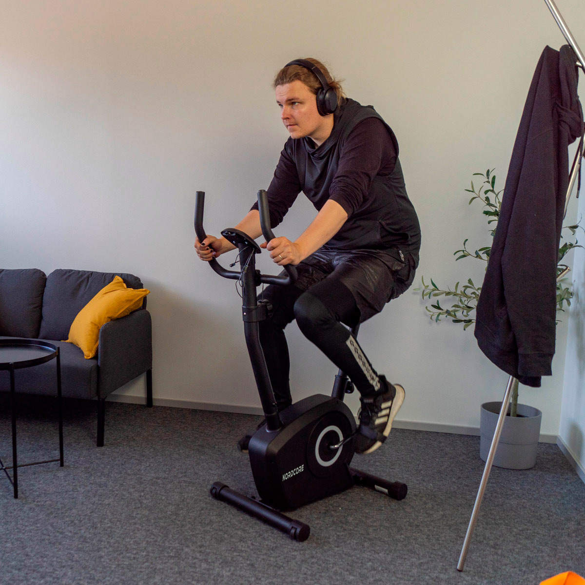 Mężczyzna na rowerze stacjonarnym, w słuchawkach. Za nim kanapa z żółtą poduszką i wieszak z czarną kurtką.