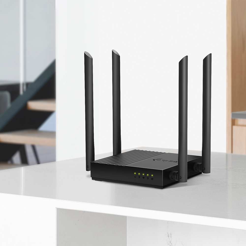 Czarny router z czterema antenami stoi na białej powierzchni. Router ma zielone światła. W tle znajdują się schody.