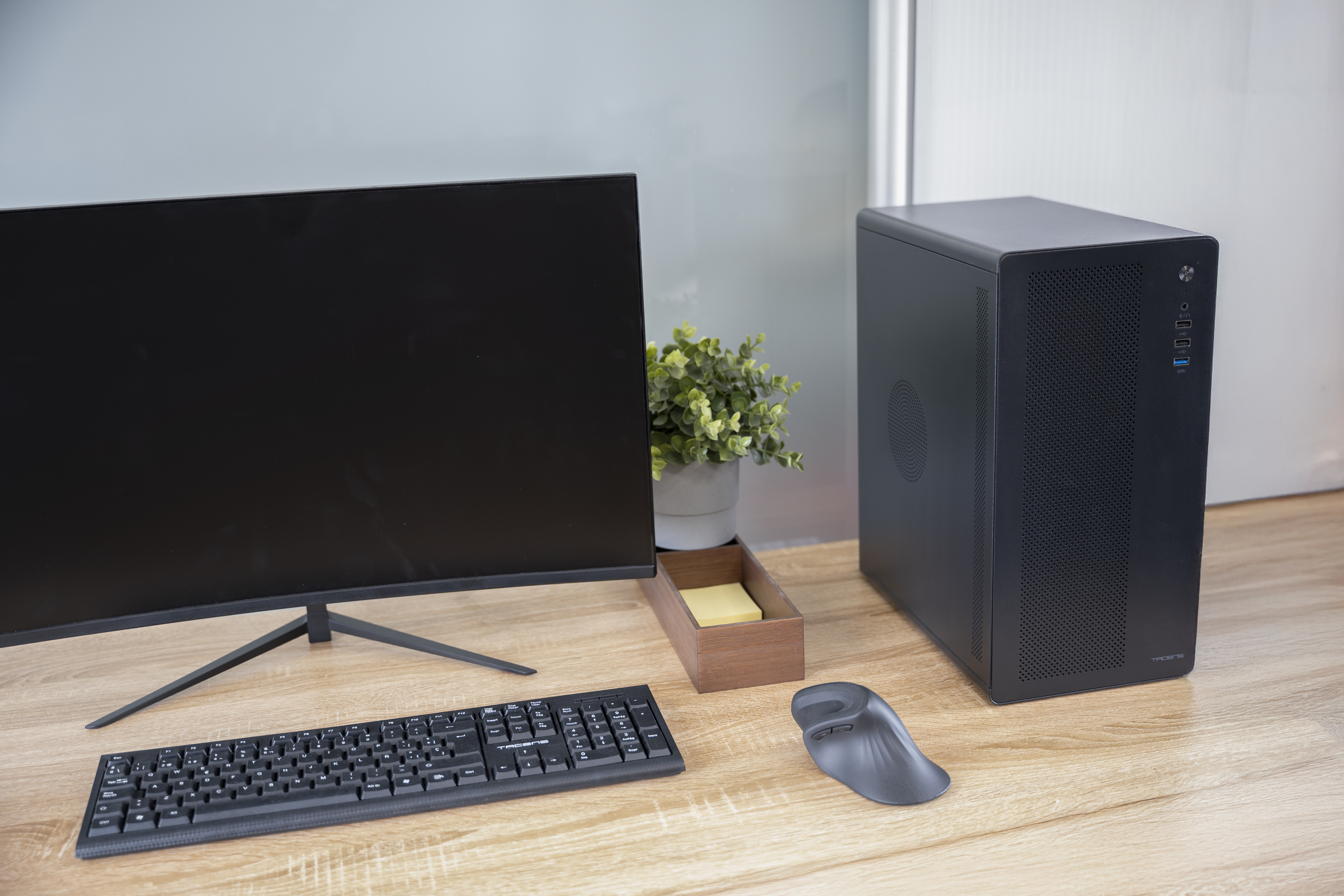 Una scrivania con monitor, tastiera, computer, mouse e pianta. La scrivania è color legno.