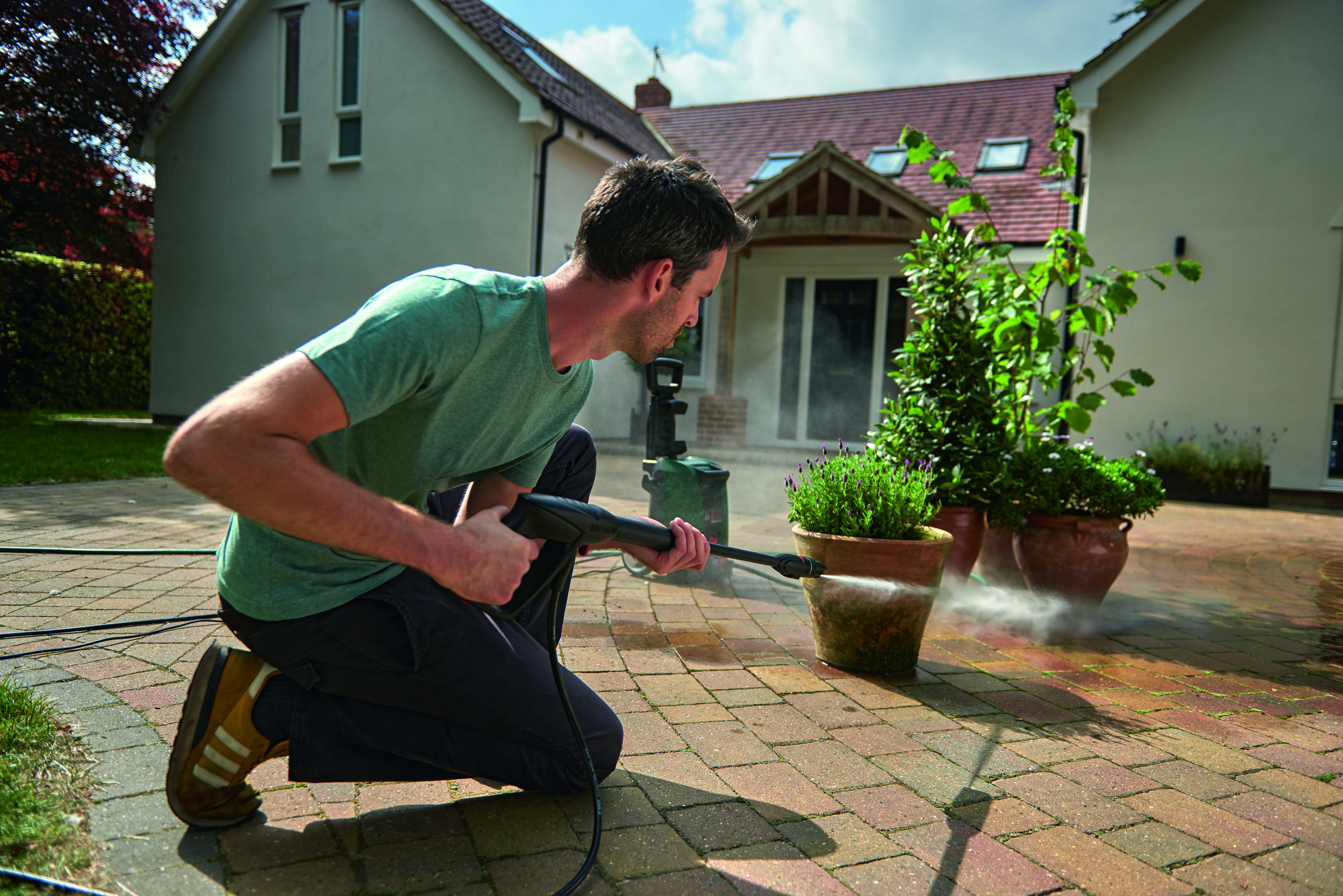 Un homme agenouillé utilise un nettoyeur haute pression sur un patio en briques. Des pots de plantes en arrière-plan.