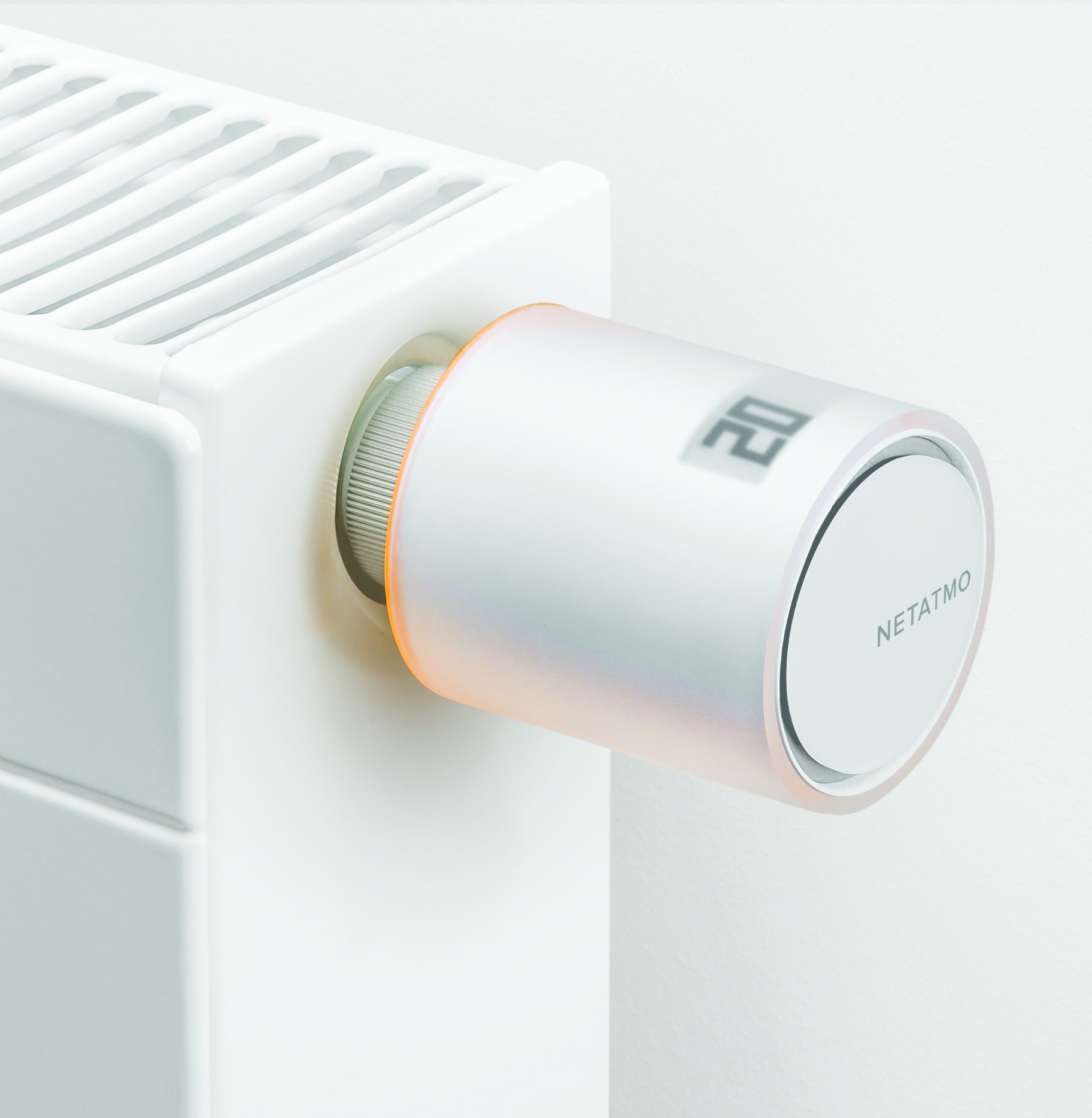 A white radiator with a Netatmo smart thermostat. The thermostat displays 20. Orange light glows around the dial.