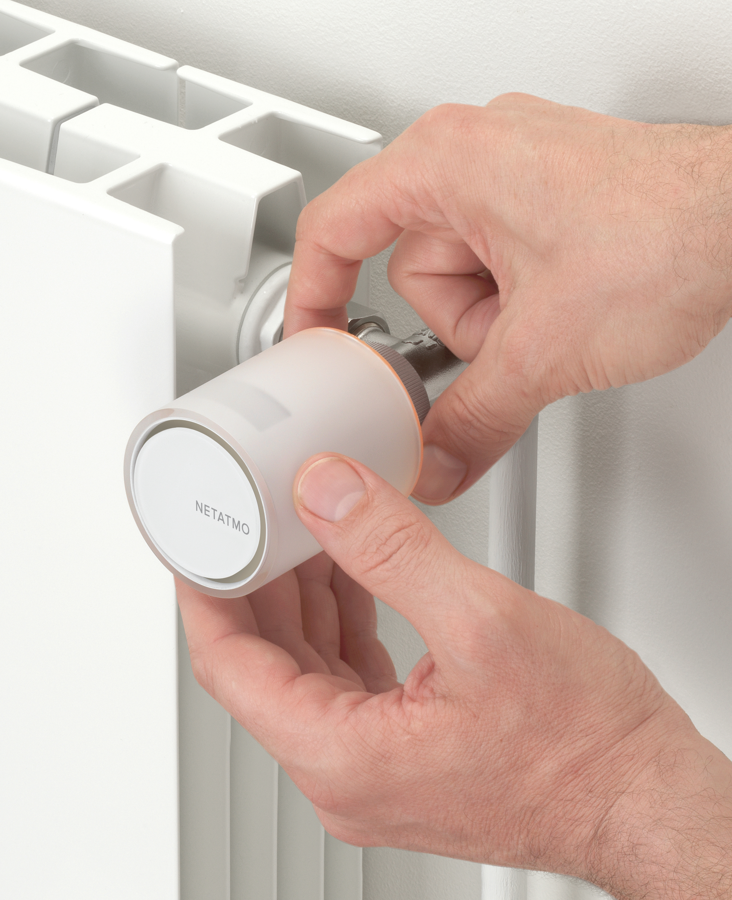 Hands adjusting a Netatmo thermostat on a white radiator. The device is white and orange, with the brand name visible.