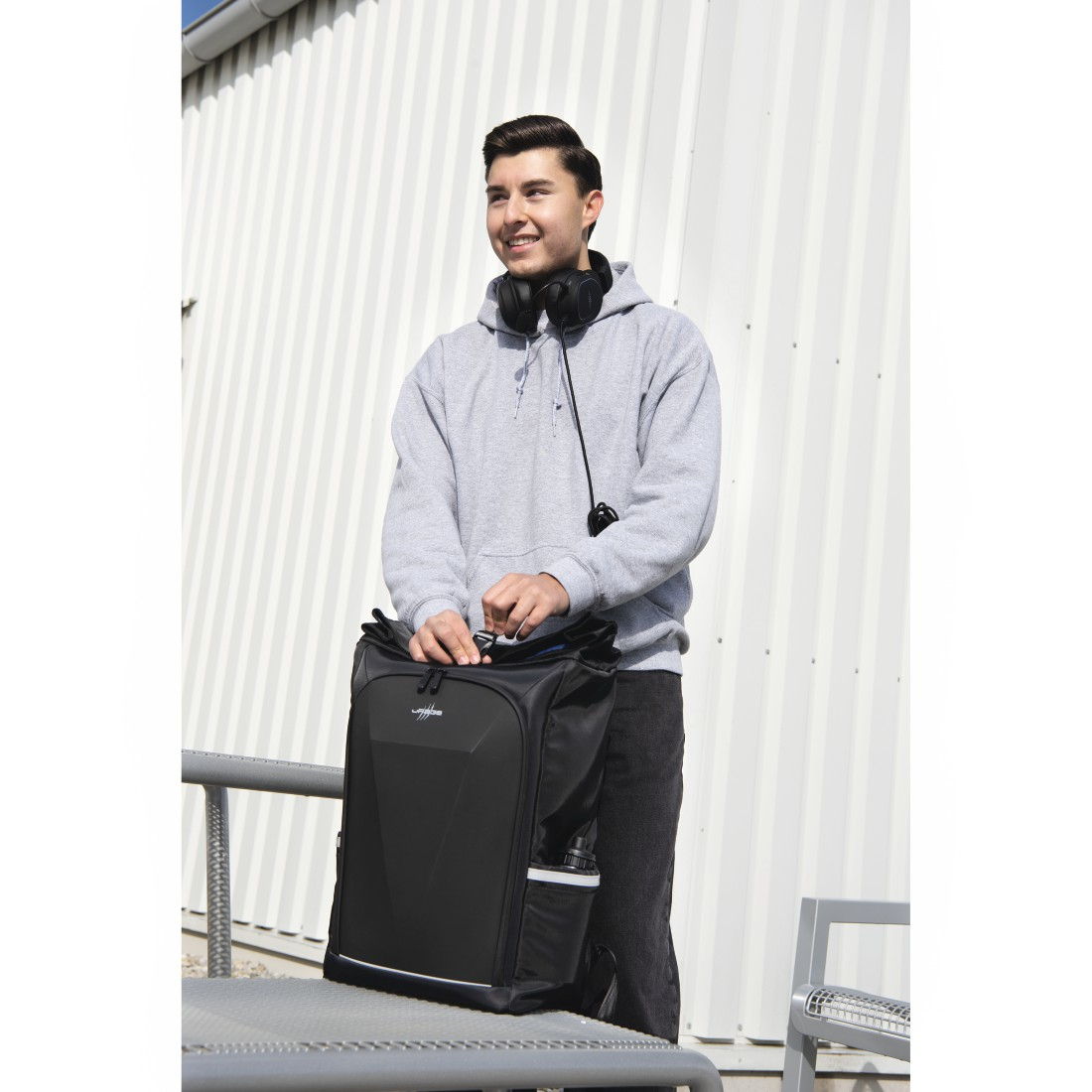 Ein Mann steht draußen und hält einen schwarzen Rucksack. Er trägt einen grauen Hoodie und Kopfhörer und lächelt. Im Hintergrund ist ein weißes Gebäude.