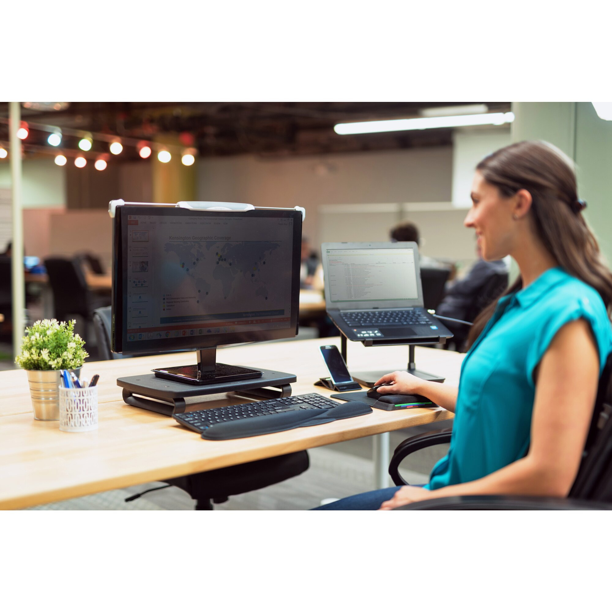 Eine Frau sitzt an einem Schreibtisch mit Computer und Laptop. Sie arbeitet und lächelt. Es gibt eine Pflanze und eine Tastatur.