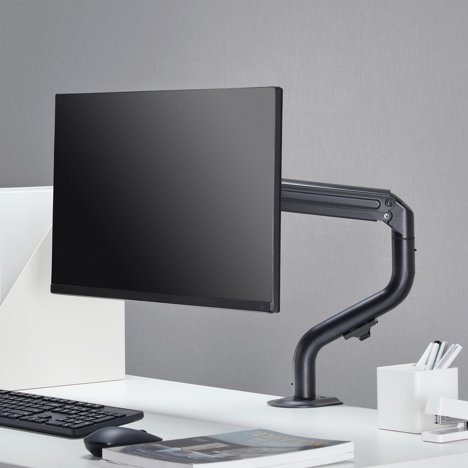 Monitor komputera jest zamontowany na czarnym ramieniu nad białym biurkiem. Obecna jest również klawiatura, mysz i inne artykuły biurowe.